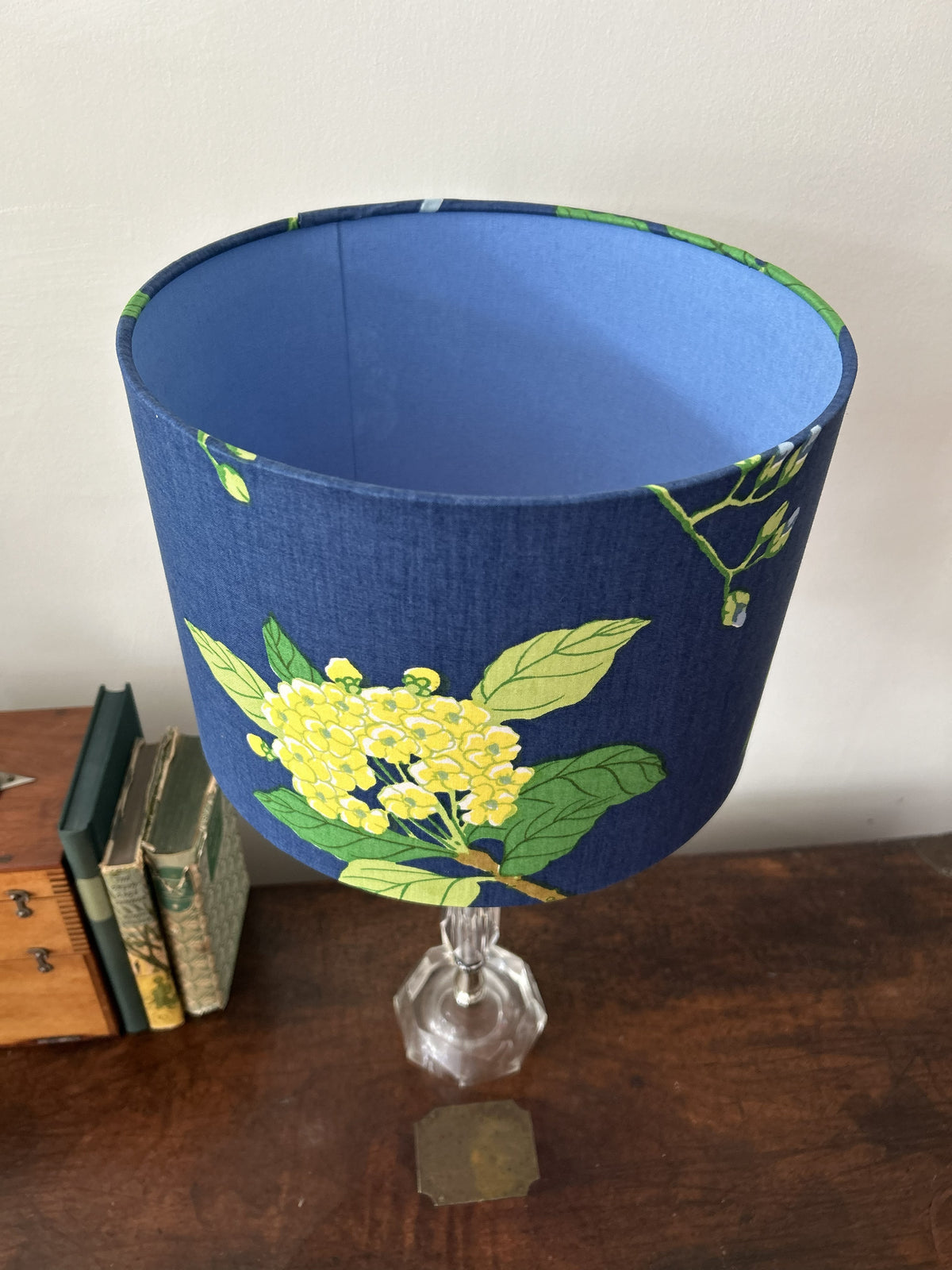 A fabric lampshade with a blue base and a yellow floral print, mounted on a clear faceted glass stem lamp. The lamp is set on a dark wooden table next to some vintage books against a plain off-white wall.