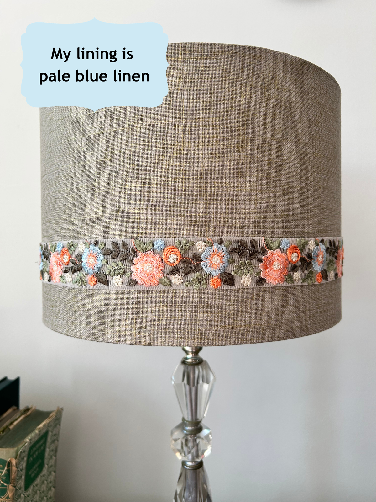 A table lamp shade made of textured beige linen with a decorative band of embroidered flowers in coral, orange, and blue-gray tones. The lamp has a clear faceted glass base.