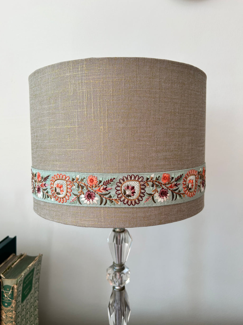 A cylindrical table lamp shade made of textured beige linen, featuring a decorative band of colorful floral embroidery around the middle. The lamp is supported by a clear faceted glass stem, set against a plain off-white wall with a stack of books partially visible on the left.