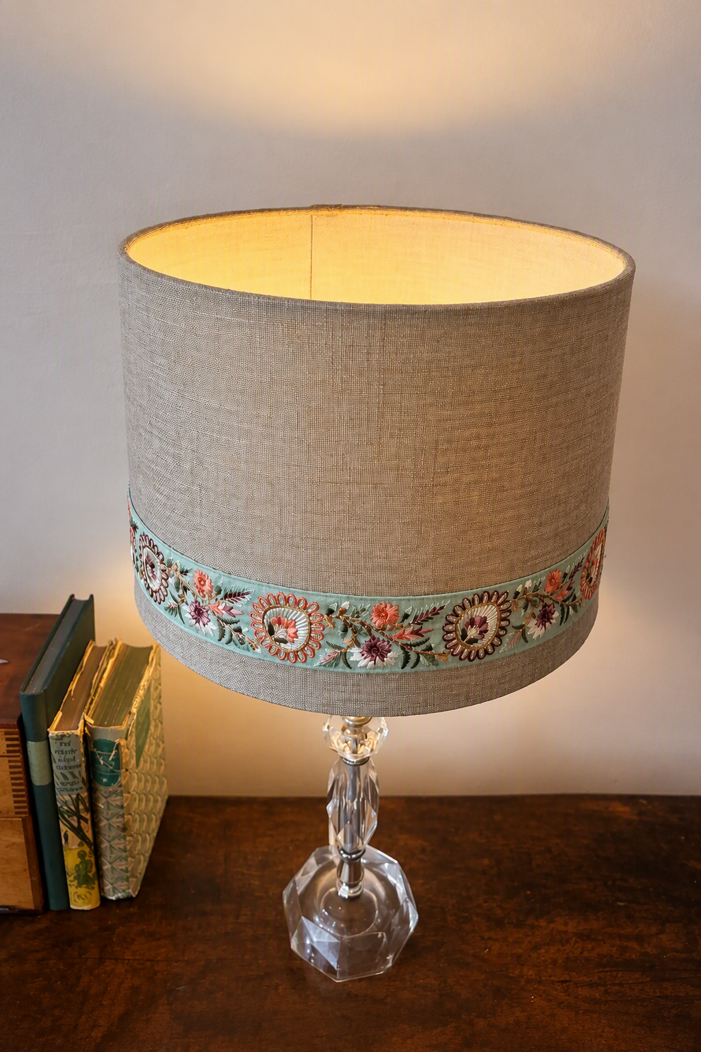 A table lamp with a beige linen drum shade featuring a decorative band of colorful floral embroidery, supported by a faceted clear glass base, set on a dark wooden table next to books.
