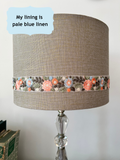 A drum shaped table lamp shade made of textured gold flecked, beige linen with a decorative band of embroidered flowers in coral, orange, and blue-grey tones. The lamp has a clear faceted glass base. There is a speech bubble over the shade that says 'my lining is pale blue linen'