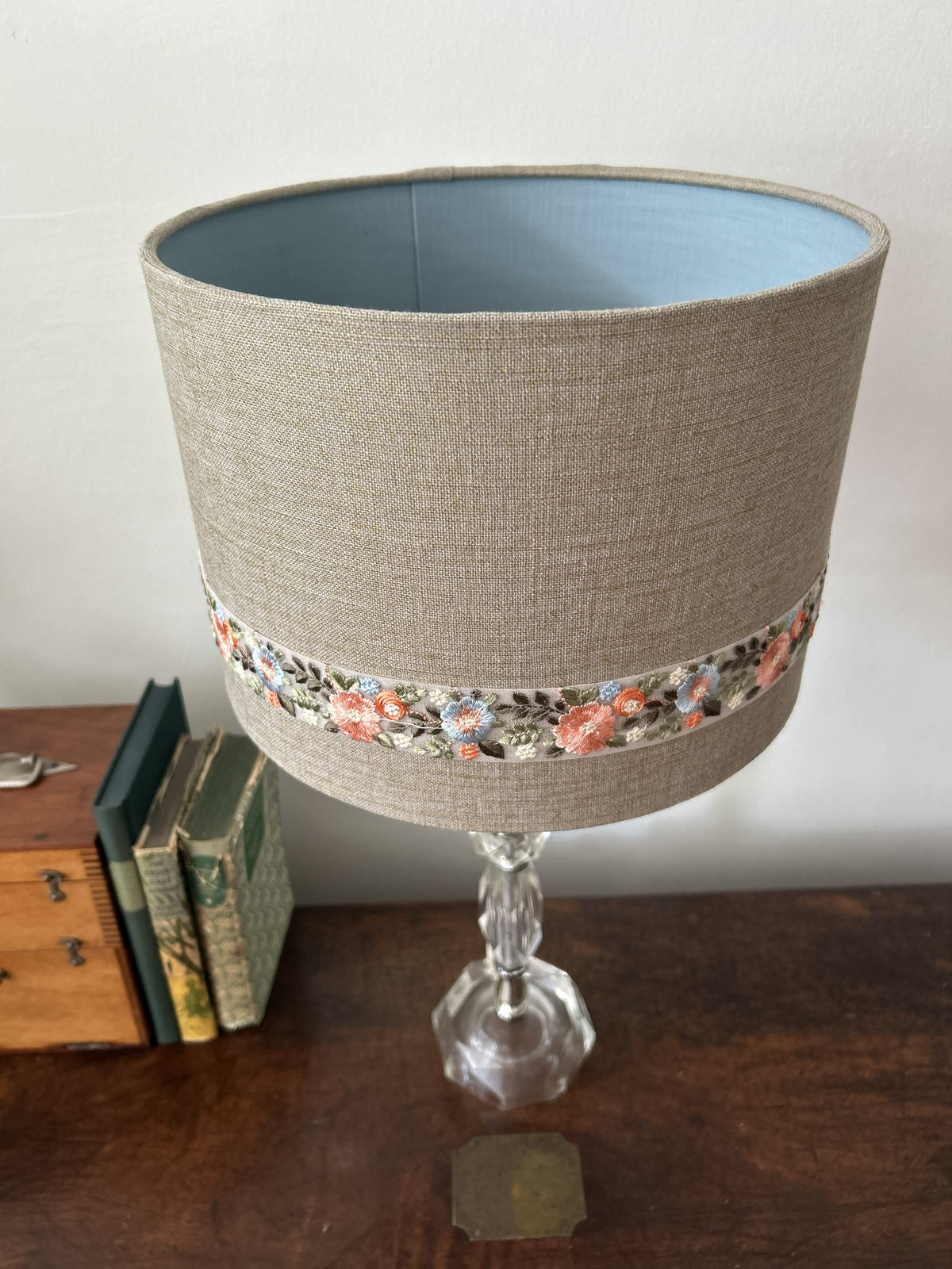 A table lamp shade made of beige linen fabric with a decorative band of colorful floral embroidery around the middle, displayed on a glass base on a wooden table.