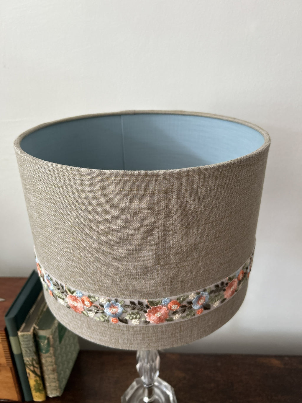 A cylindrical fabric lampshade with a textured beige linen exterior, a light blue interior, and a decorative band of small floral embroidery around the lower middle. The lampshade is shown on a glass table base next to some books against a plain wall.