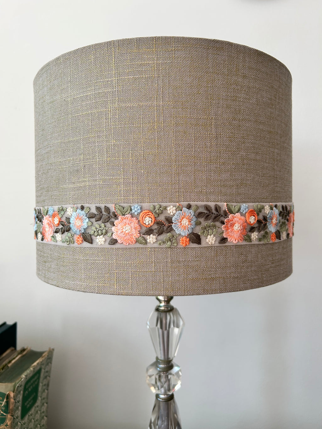 A table lamp with a cylindrical beige linen shade featuring a decorative band of embroidered flowers in coral, orange, blue, and green, mounted on a clear glass and silver-toned metal lamp base.