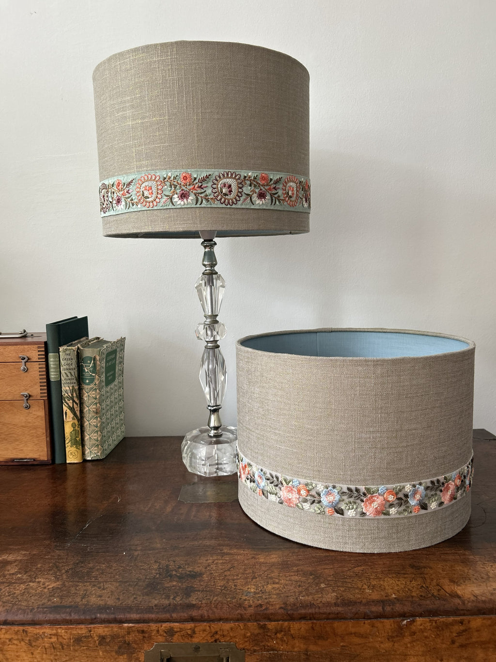 A cylindrical table lamp shade and matching lamp base displayed on a wooden table against a plain wall. The lampshade and base are upholstered in a warm beige linen fabric with a decorative band of floral embroidery around the middle.