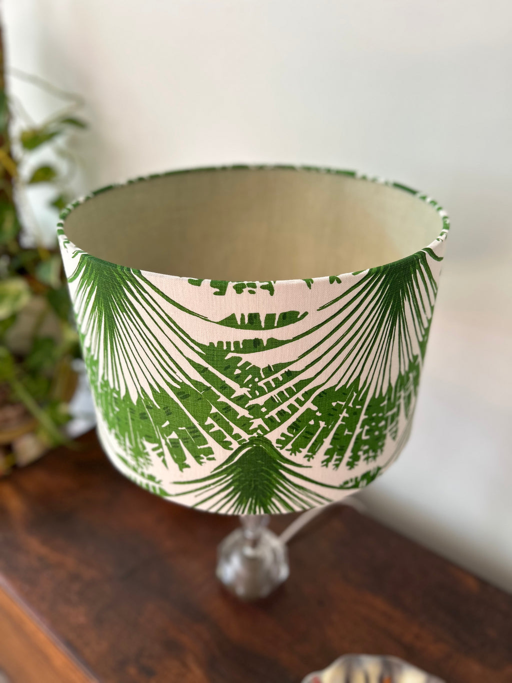 A fabric lampshade with a green palm leaf pattern on a cream background, displayed on a glass stand over a dark wooden surface.