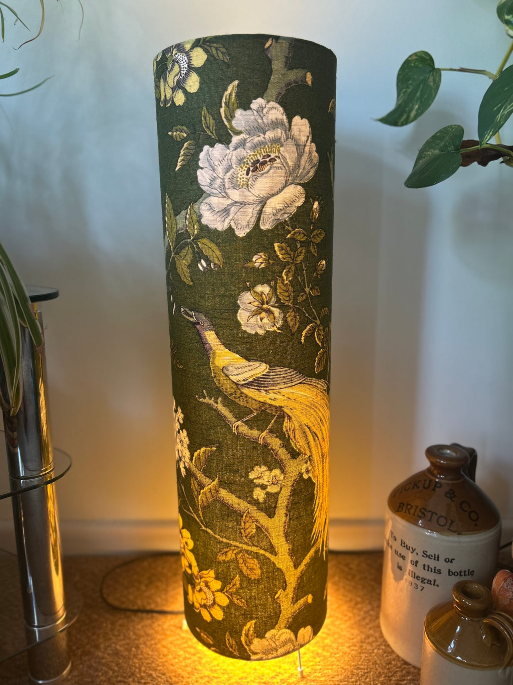 A tall cylindrical floor lamp with a dark green fabric shade featuring a vintage floral and peacock and small bird pattern, illuminated from within to cast a warm yellow glow on the floor. The lamp is styled in a home setting next to ceramic jugs and a houseplant.