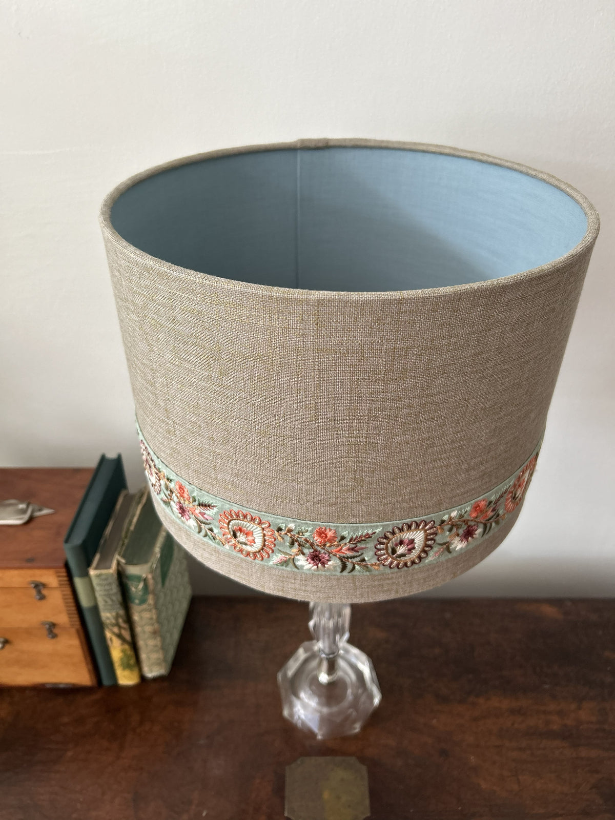 A table lamp shade made of beige gold fleck linen fabric with a decorative band of colourful floral embroidery around the lower middle, shown on a glass pedestal lamp. The lamp is placed on a dark wooden table next to some vintage books against a plain white wall.