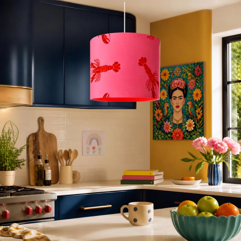 A bright pink and orange cylindrical kitchen pendant light with red lobster prints hangs in a modern kitchen with navy cabinets, a yellow accent wall, and colourful decor.