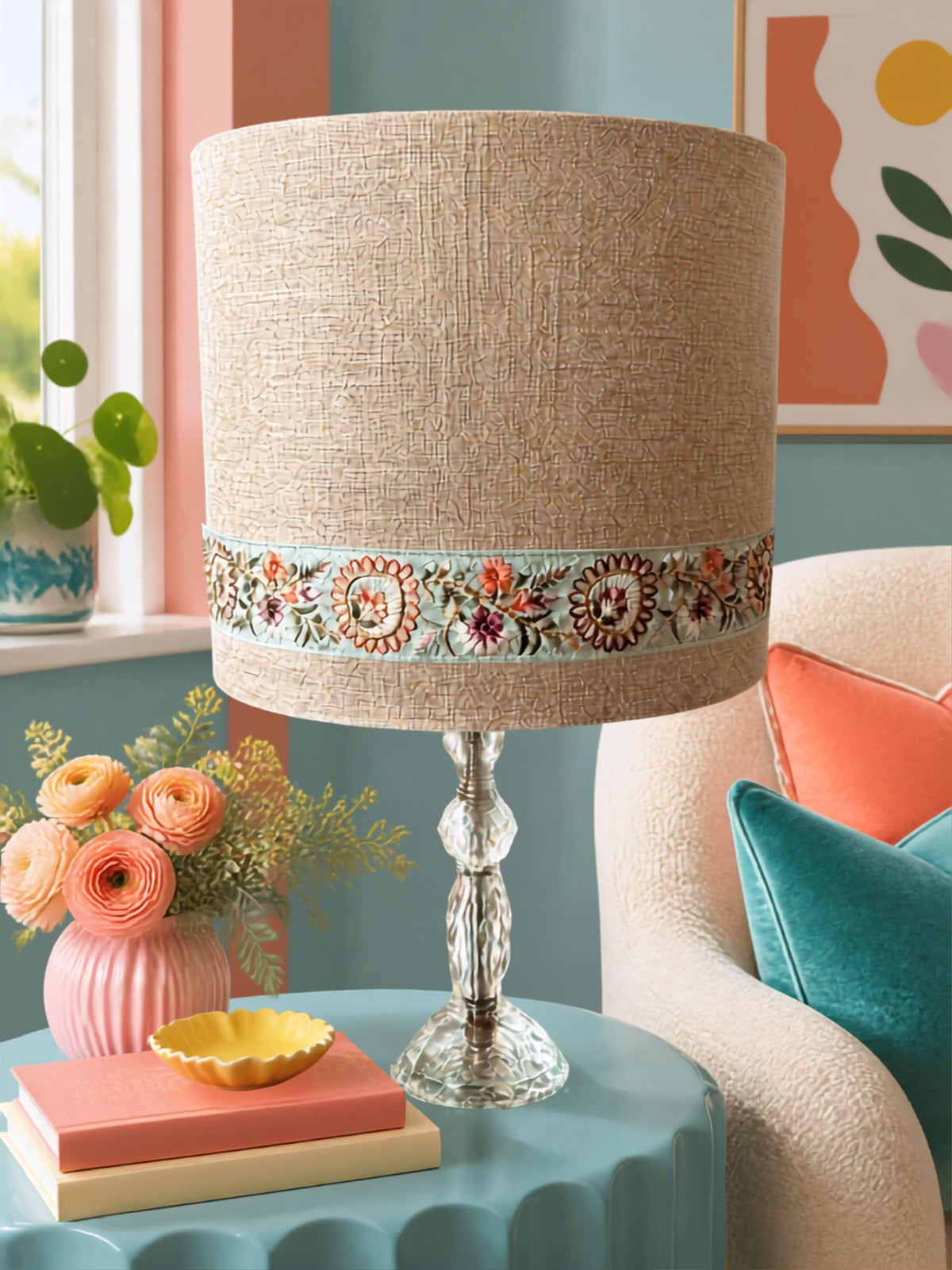 A table lamp with a golden beige linen drum shade featuring a colourful floral embroidered band around the middle, set on a turquoise side table in a styled living room.