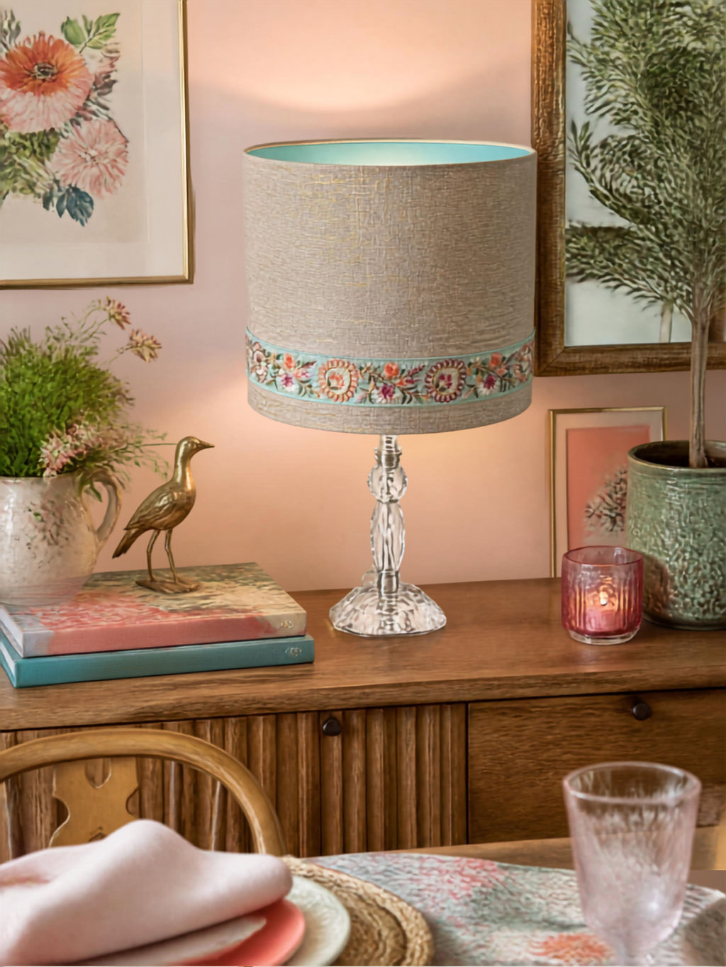 A table lamp with a textured gold beige linen drum shade, a turquoise blue inner lining, and a decorative band of colourful floral embroidery around the middle, set on a crystal-style base on a wooden sideboard.