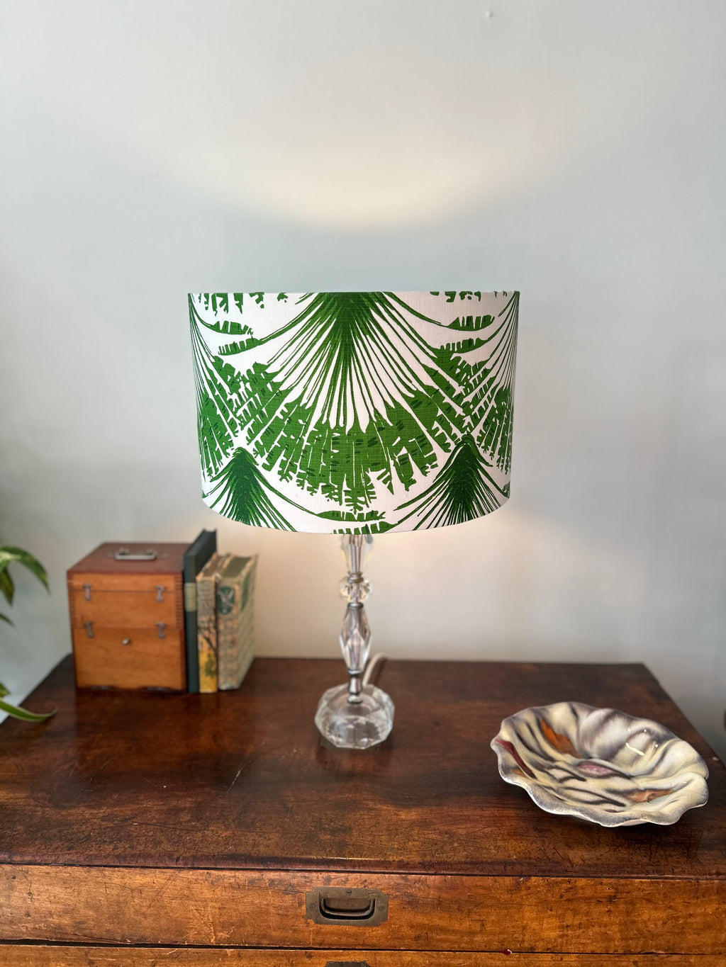 A table lamp with a white drum shade featuring a green palm frond pattern, set on a dark wooden chest next to a small wooden box and a ceramic dish against a plain wall.