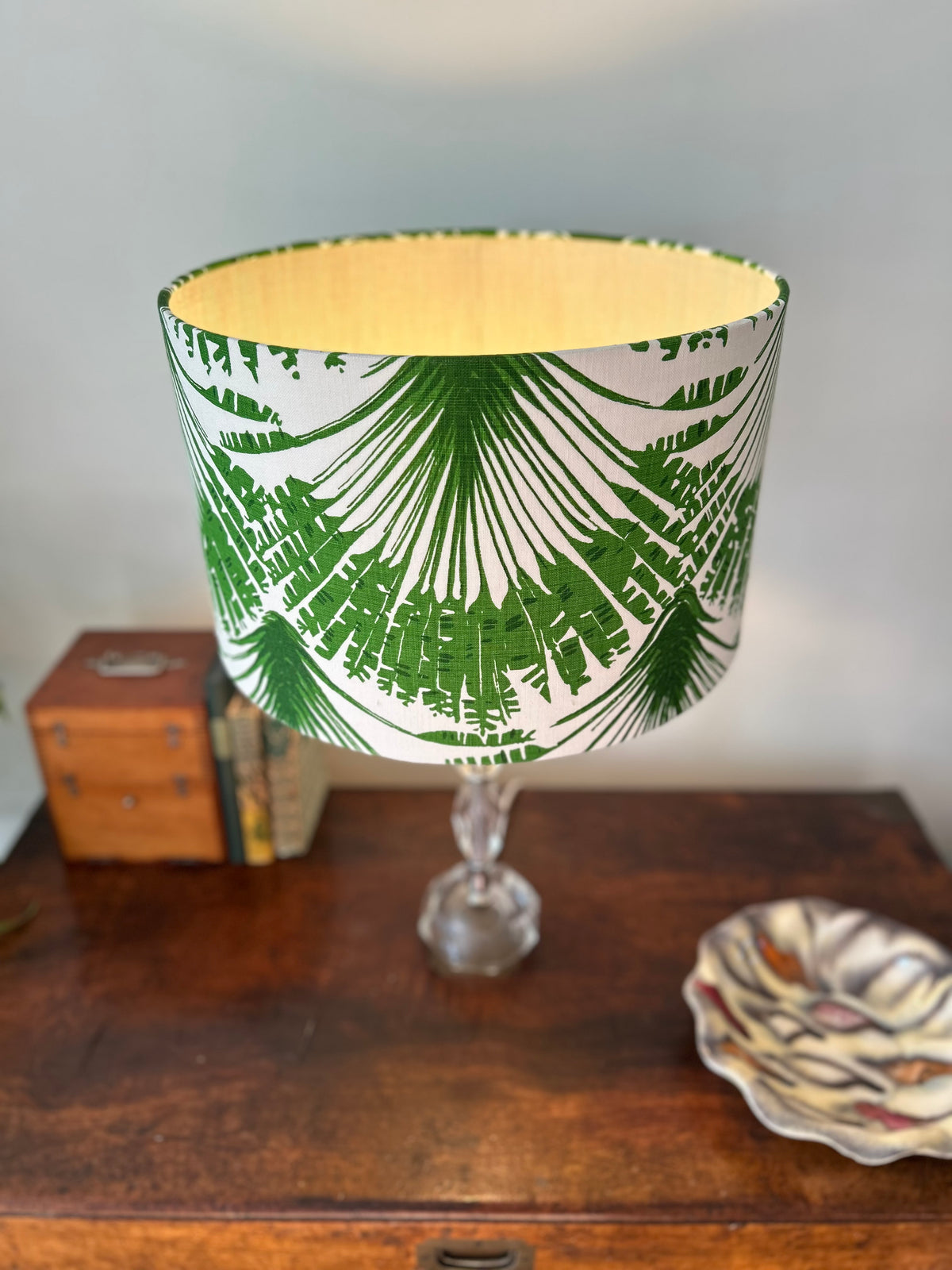 A table lamp with a white drum shade featuring a green palm leaf pattern and a green heavy cotton interior, mounted on a clear glass stem base and set on a wooden table.