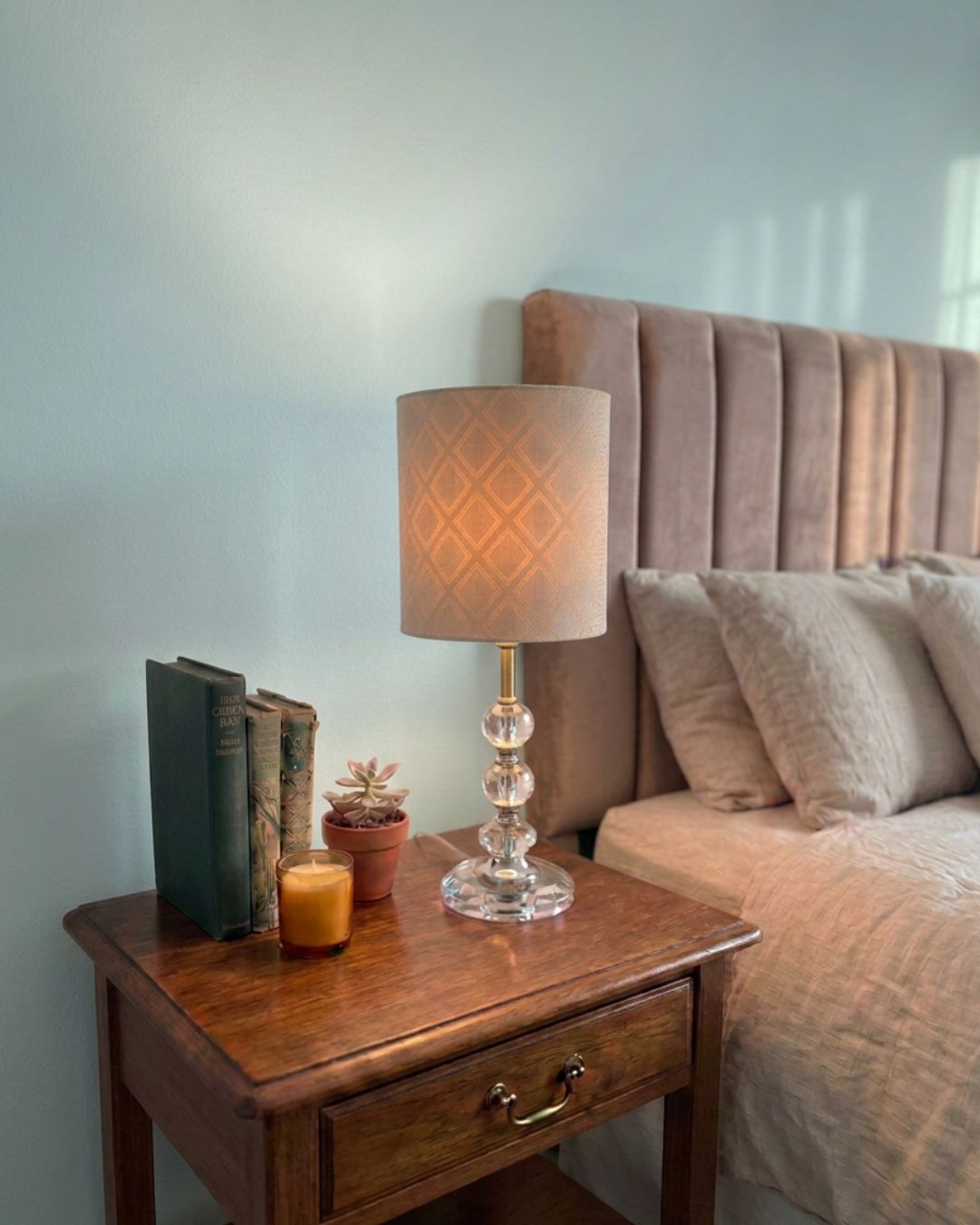 A bedside table in a bedroom setting with a table lamp featuring a beige slub satin drum shade with the gemoetric Liberty Terrace pattern visible through the satin as the light is on, and clear glass base, next to a bed with a tufted headboard.