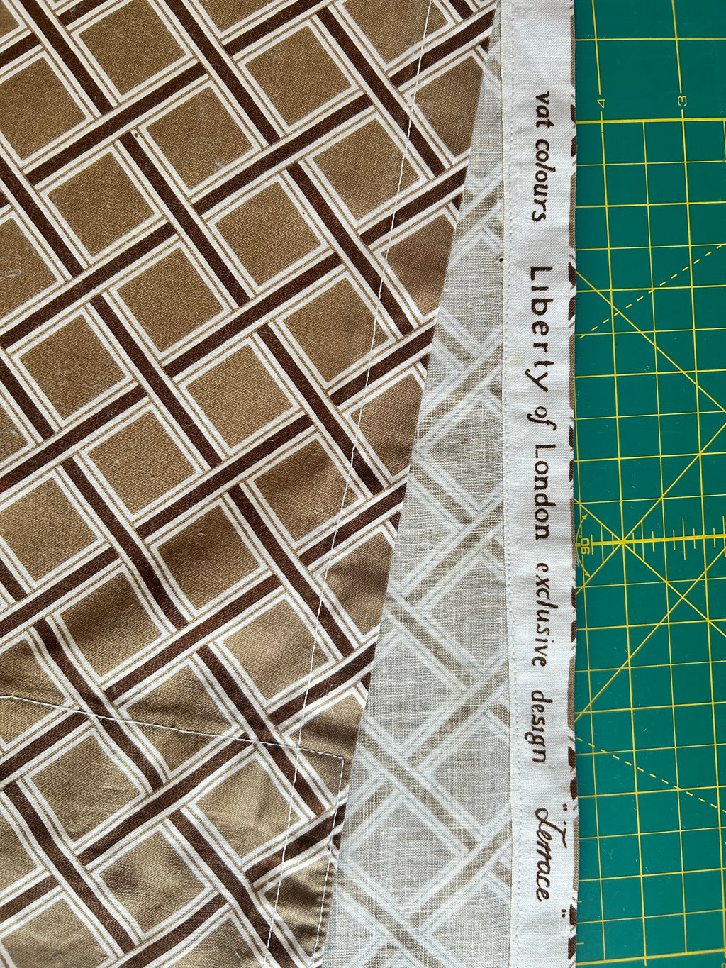 A close-up of patterned fabric with a brown and beige geometric lattice design, a white selvedge edge printed with 'Liberty of London exclusive design Terrace', and a green cutting mat visible on the right.