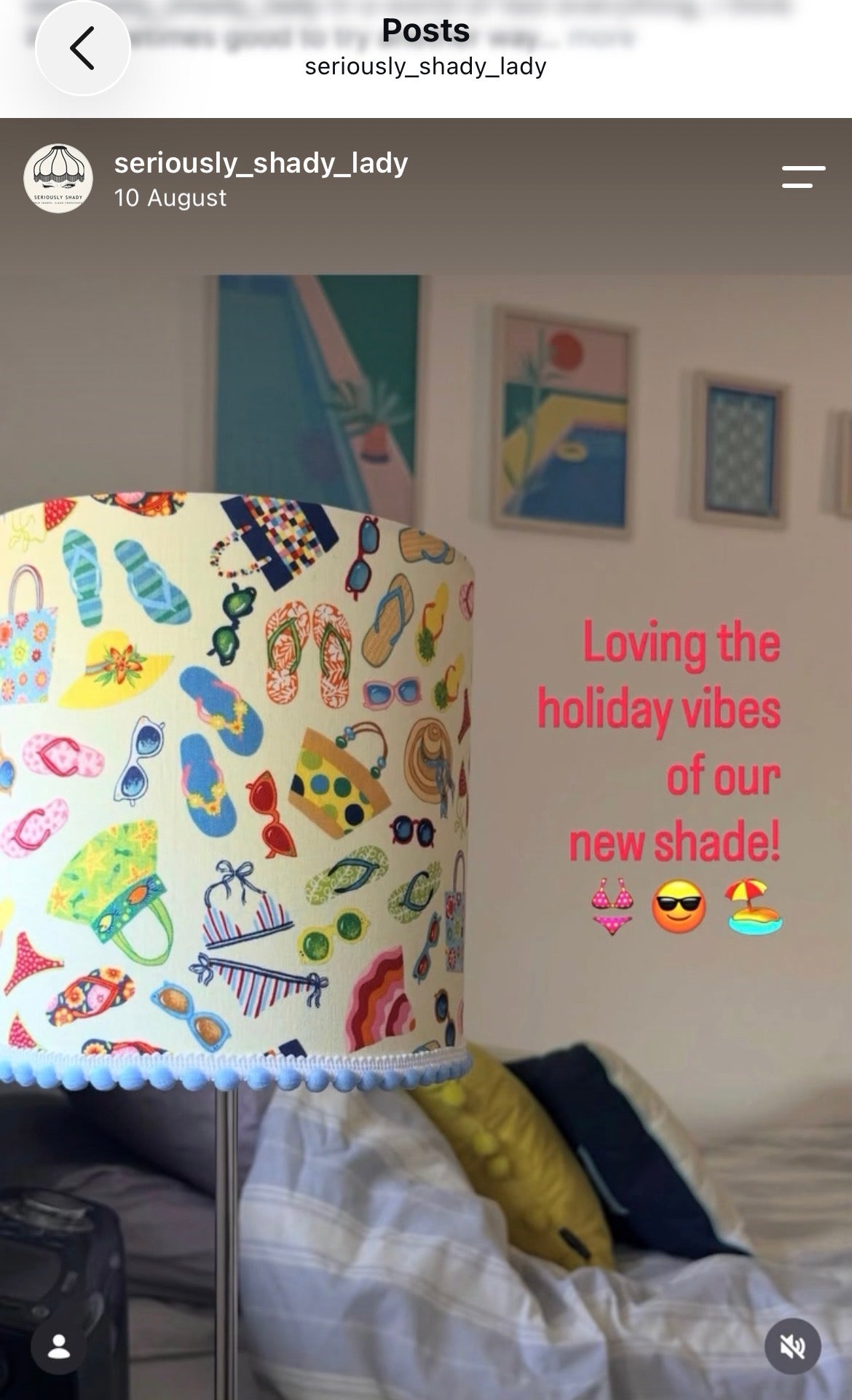 A lamp shade with a colorful beach-themed print of flip-flops, sunglasses, hats, and umbrellas, shown in a bedroom setting with a bed and framed art in the background. Pink text on the right reads, 'Loving the holiday vibes of our new shade!'.
