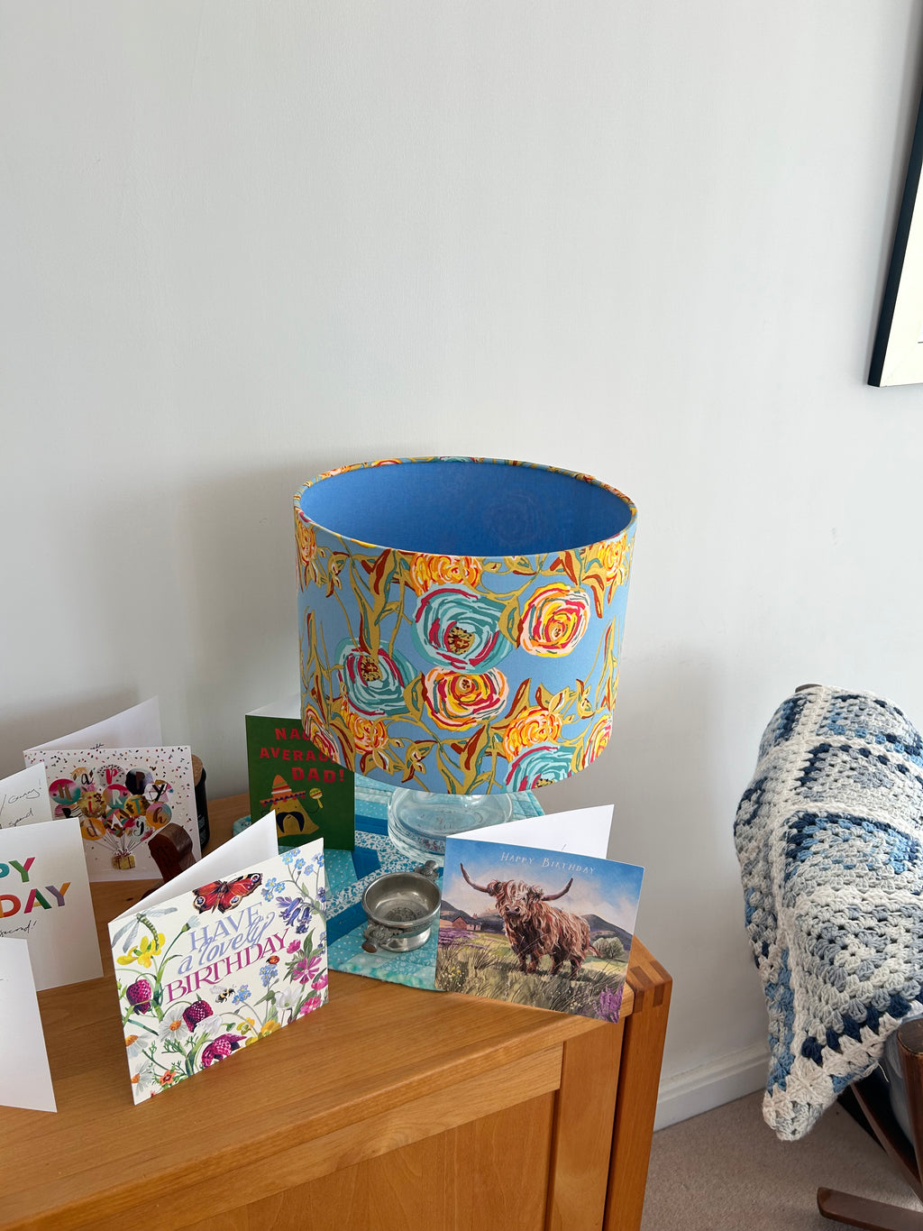 A decorative lampshade with a colorful floral pattern in yellow, orange, and blue, set on a wooden side table next to greeting cards and a crocheted throw blanket.