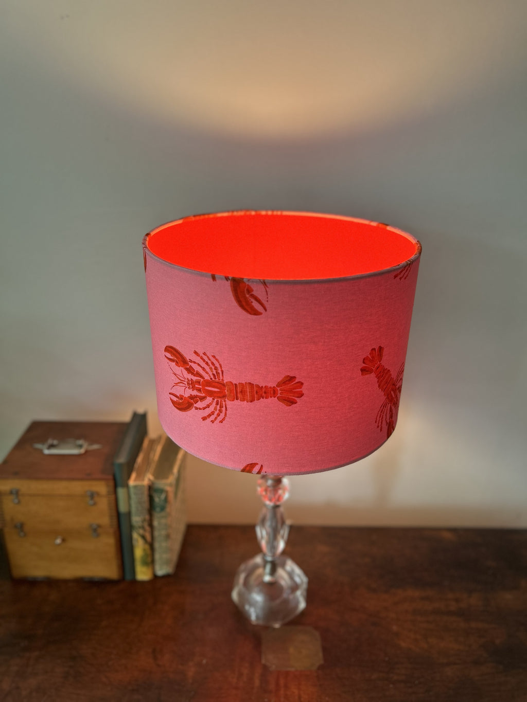 A pink fabric table lampshade with a red lobster pattern, mounted on a clear glass stem base and set on a wooden table next to a small wooden box.