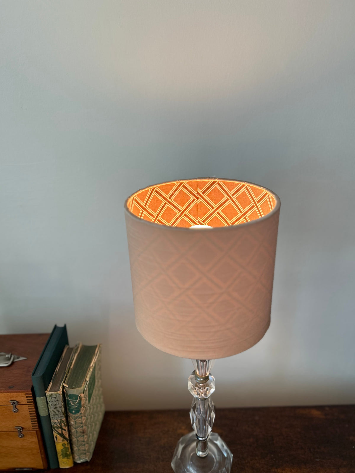 A table lamp with a beige textured drum shade, featuring an brown and beige diamond pattern on the inner lining, mounted on a clear glass stem base and set on a wooden surface.