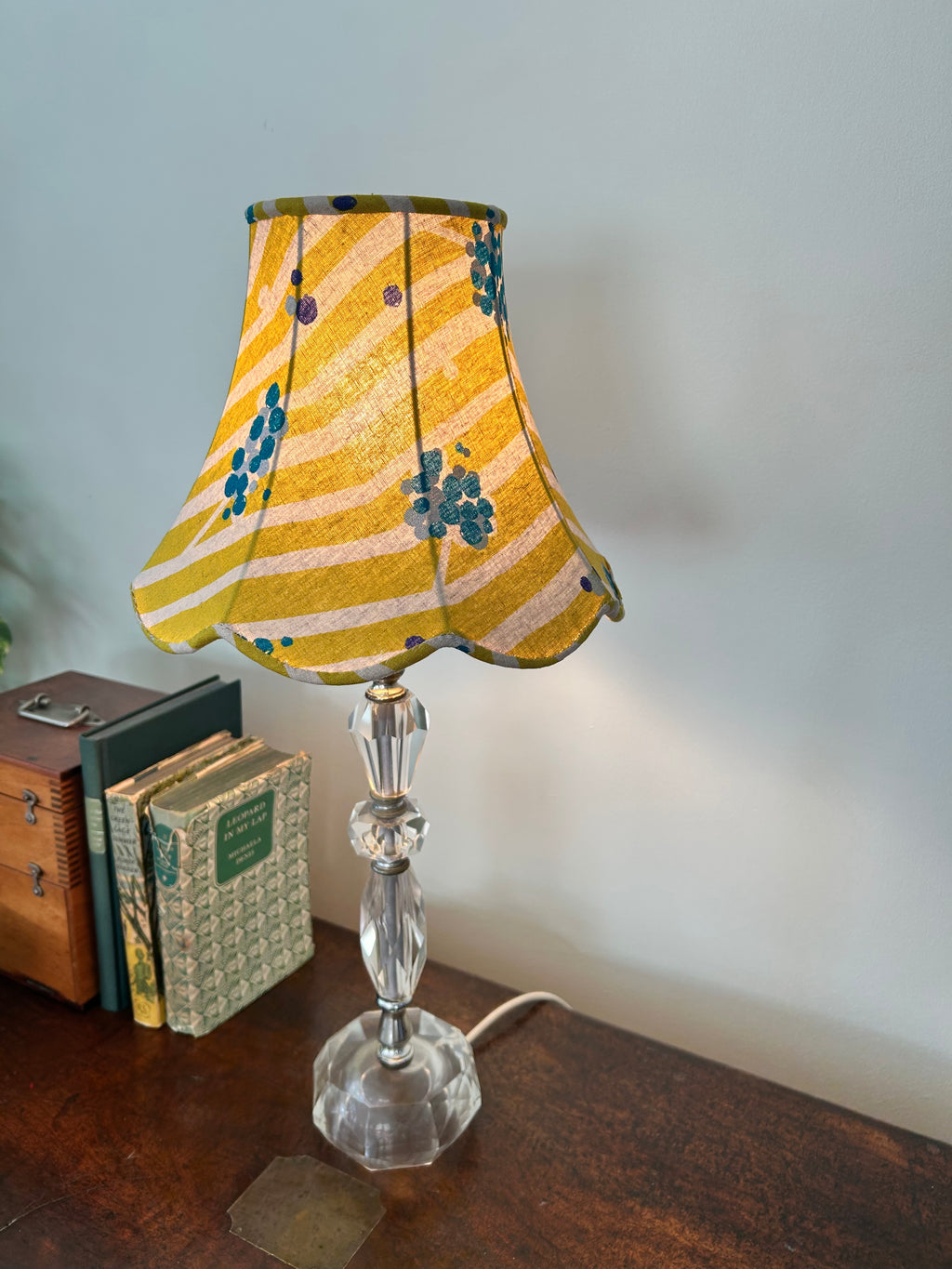 Lit lime-green empire lampshade with cream stripes, teal dots and a scalloped edge, displayed on a clear glass base.