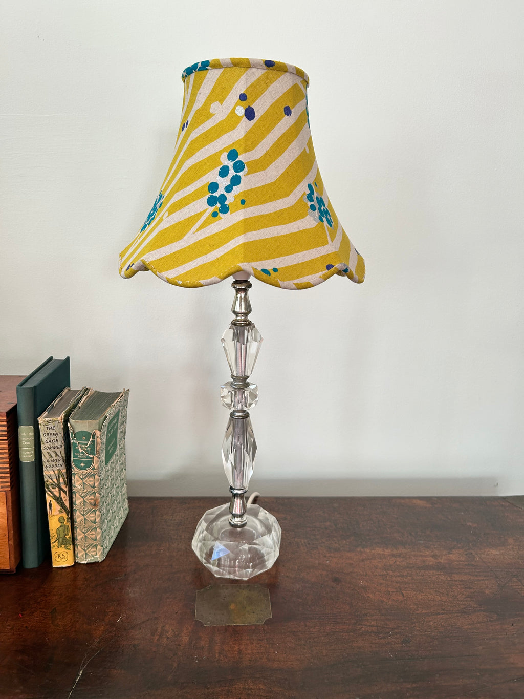 Unlit lime-green striped empire lampshade with teal dots and a scalloped edge, on a faceted glass base beside books.
