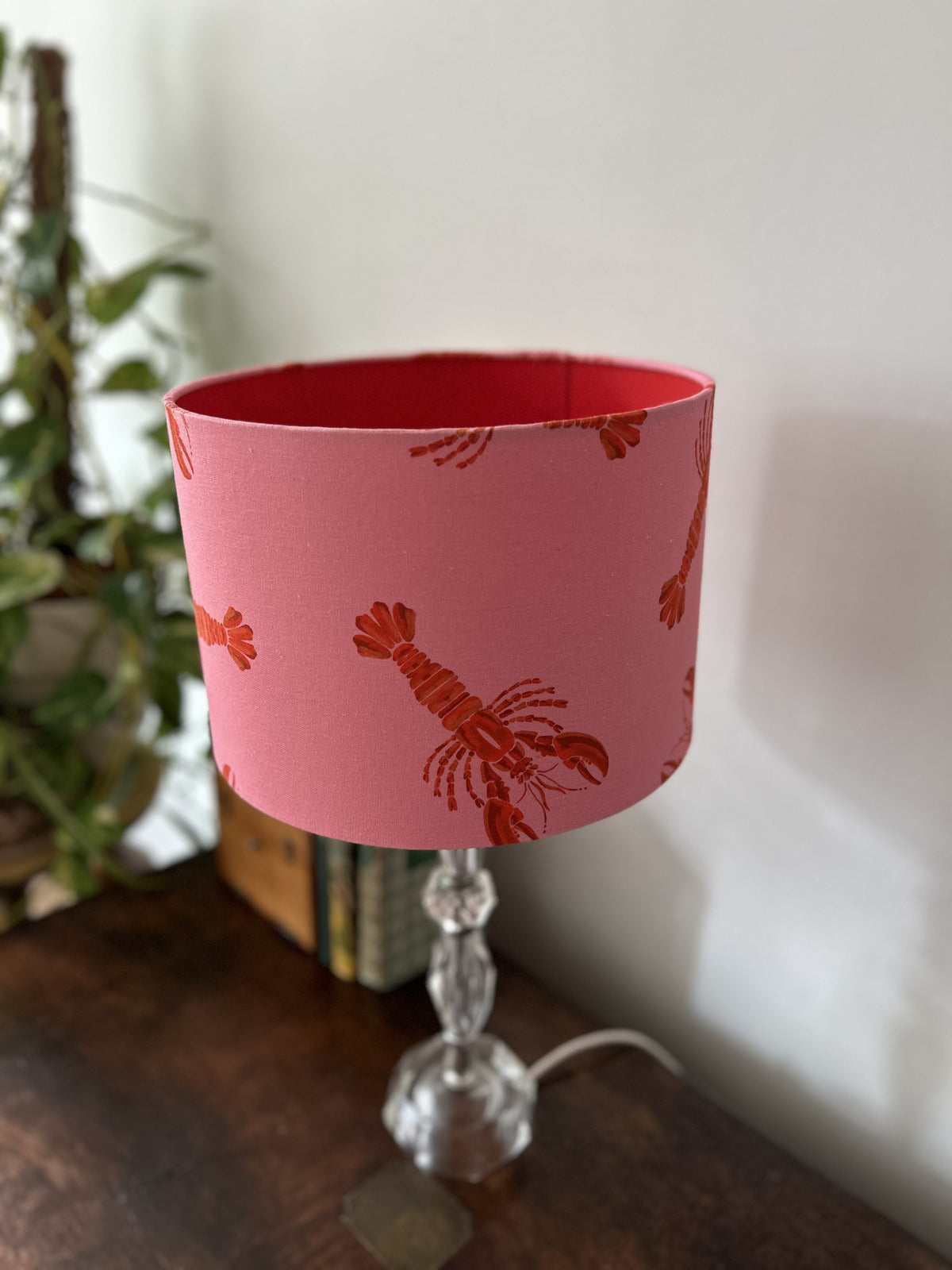 A pink table lamp shade with a red lobster pattern, mounted on a clear glass stem lamp on a dark wooden table against a white wall.