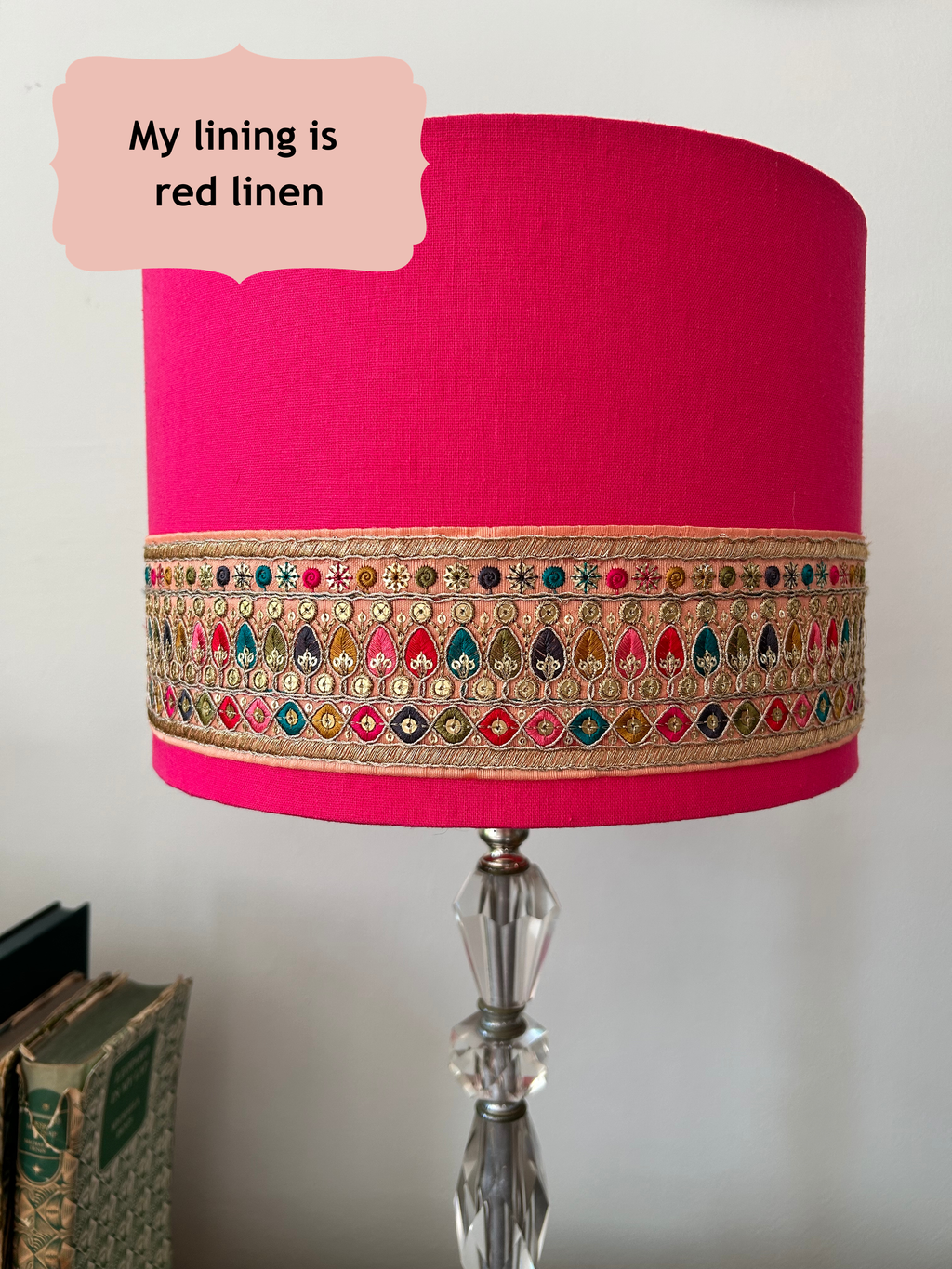 A cylindrical table lamp shade made of bright pink linen fabric, decorated with a wide gold metallic embroidered band featuring colourful thread details. The lamp shade is mounted on a clear faceted glass lamp stand, with a text bubble in the top left reading 'My lining is red linen'.