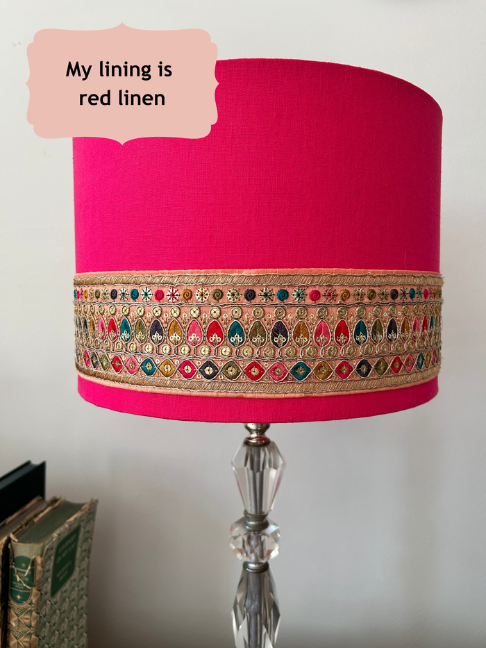 A cylindrical table lamp shade made of bright pink linen fabric, decorated with a wide gold metallic embroidered band featuring colourful thread details. The lamp shade is mounted on a clear faceted glass lamp stand, with a text bubble in the top left reading 'My lining is red linen'.
