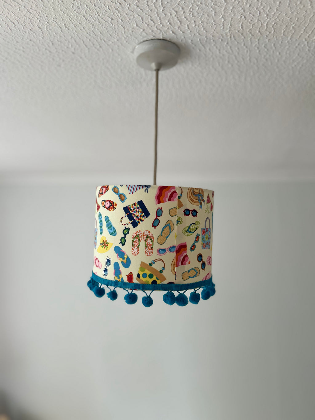 A cylindrical pendant lampshade with a colorful beach-themed pattern of flip-flops, umbrellas, and sunglasses, hanging from a silver cord on a white ceiling.