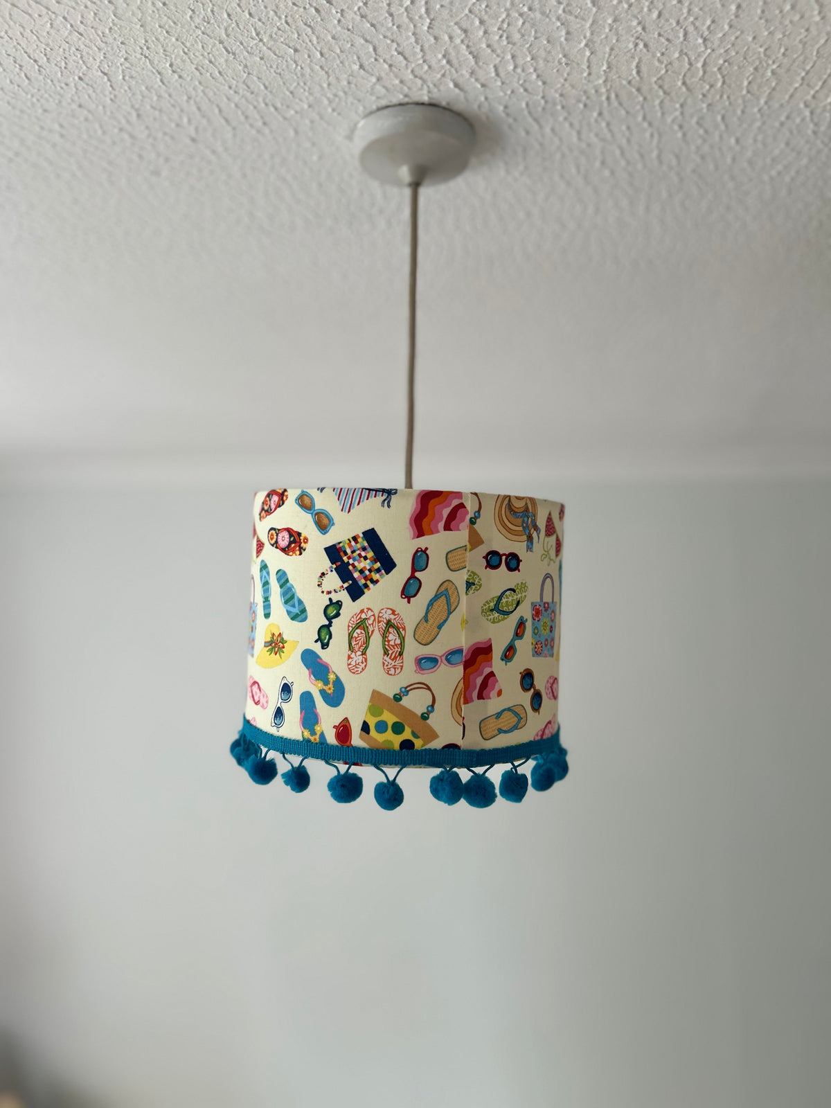 A cylindrical pendant lampshade with a colorful beach-themed pattern of flip-flops, umbrellas, and sunglasses, hanging from a silver cord on a white ceiling.