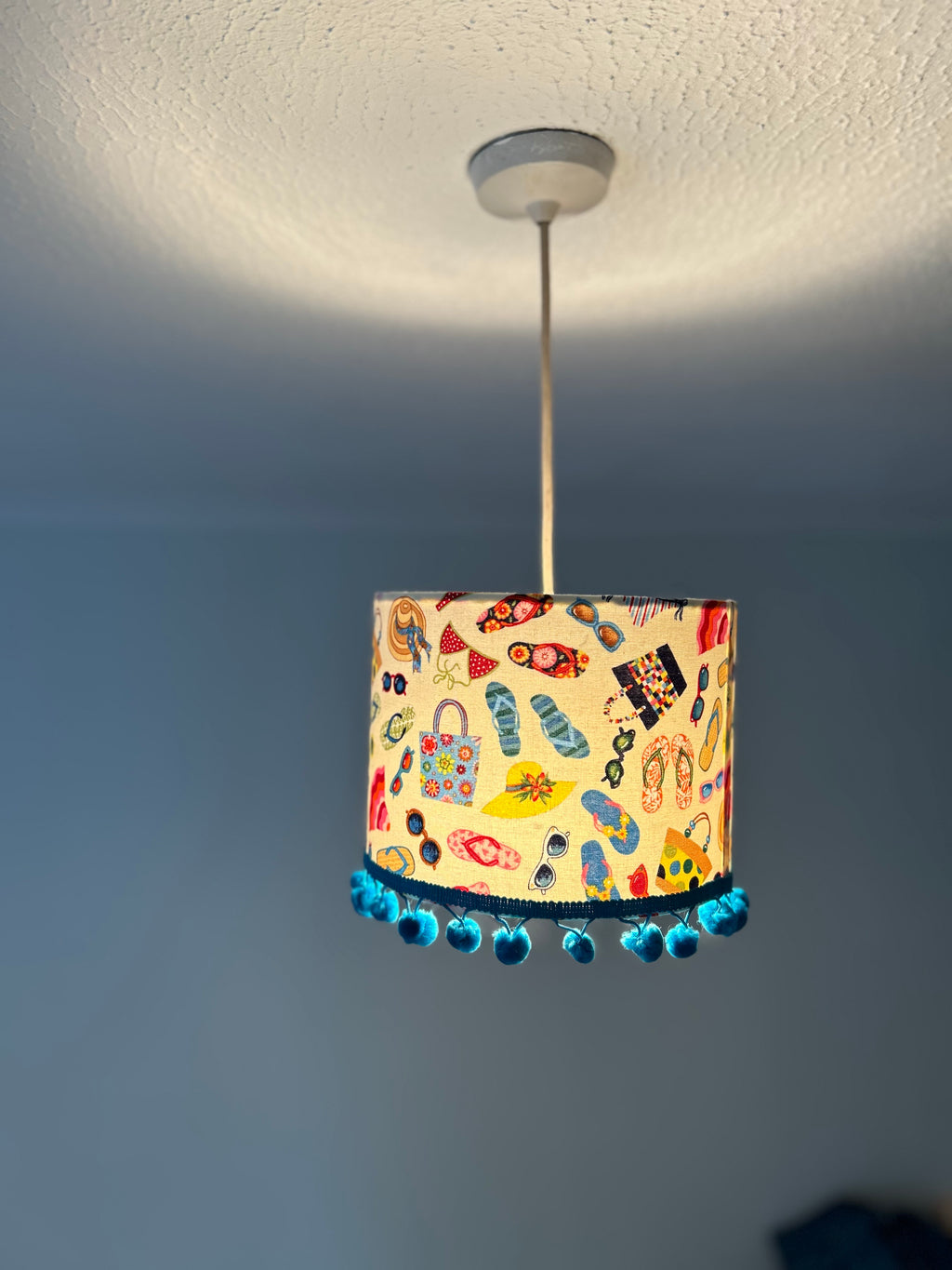 A cylindrical pendant lampshade with a colorful beach-themed print of flip-flops, sunglasses, hats, and handbags, finished with a blue beaded fringe, hanging from a ceiling in a room.