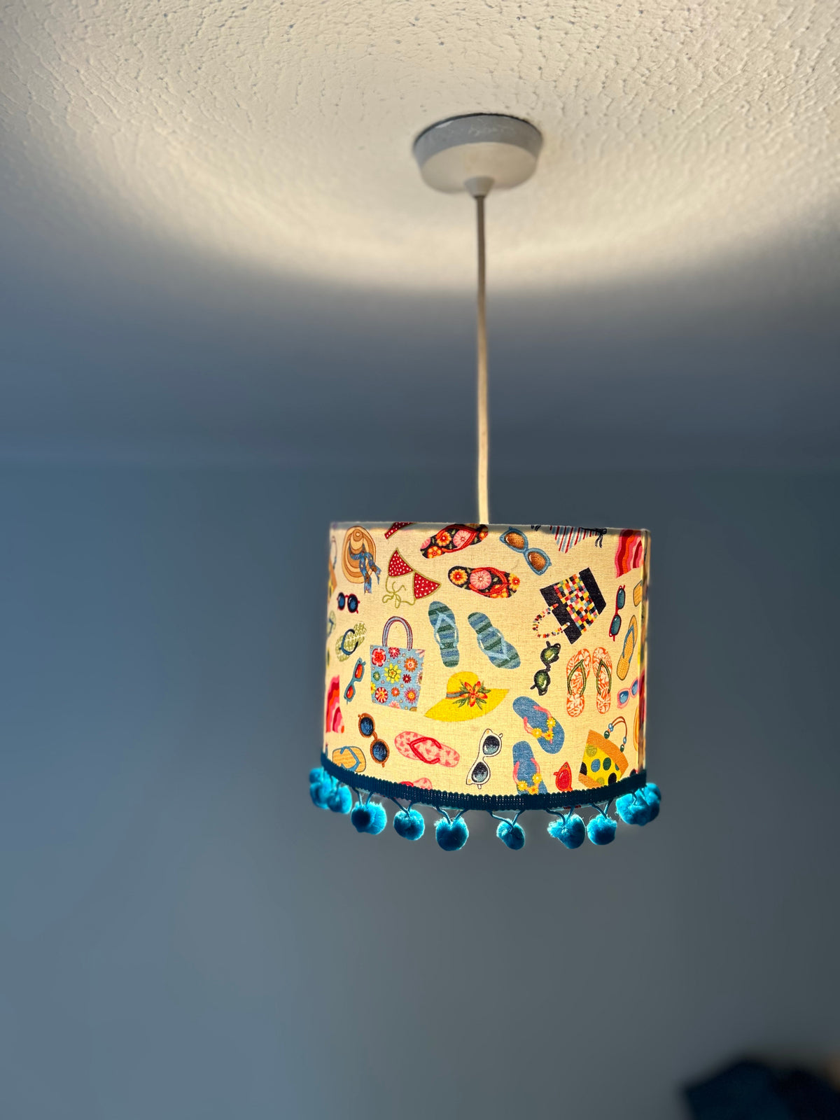 A cylindrical pendant lampshade with a colorful beach-themed print of flip-flops, sunglasses, hats, and handbags, finished with a blue beaded fringe, hanging from a ceiling in a room.