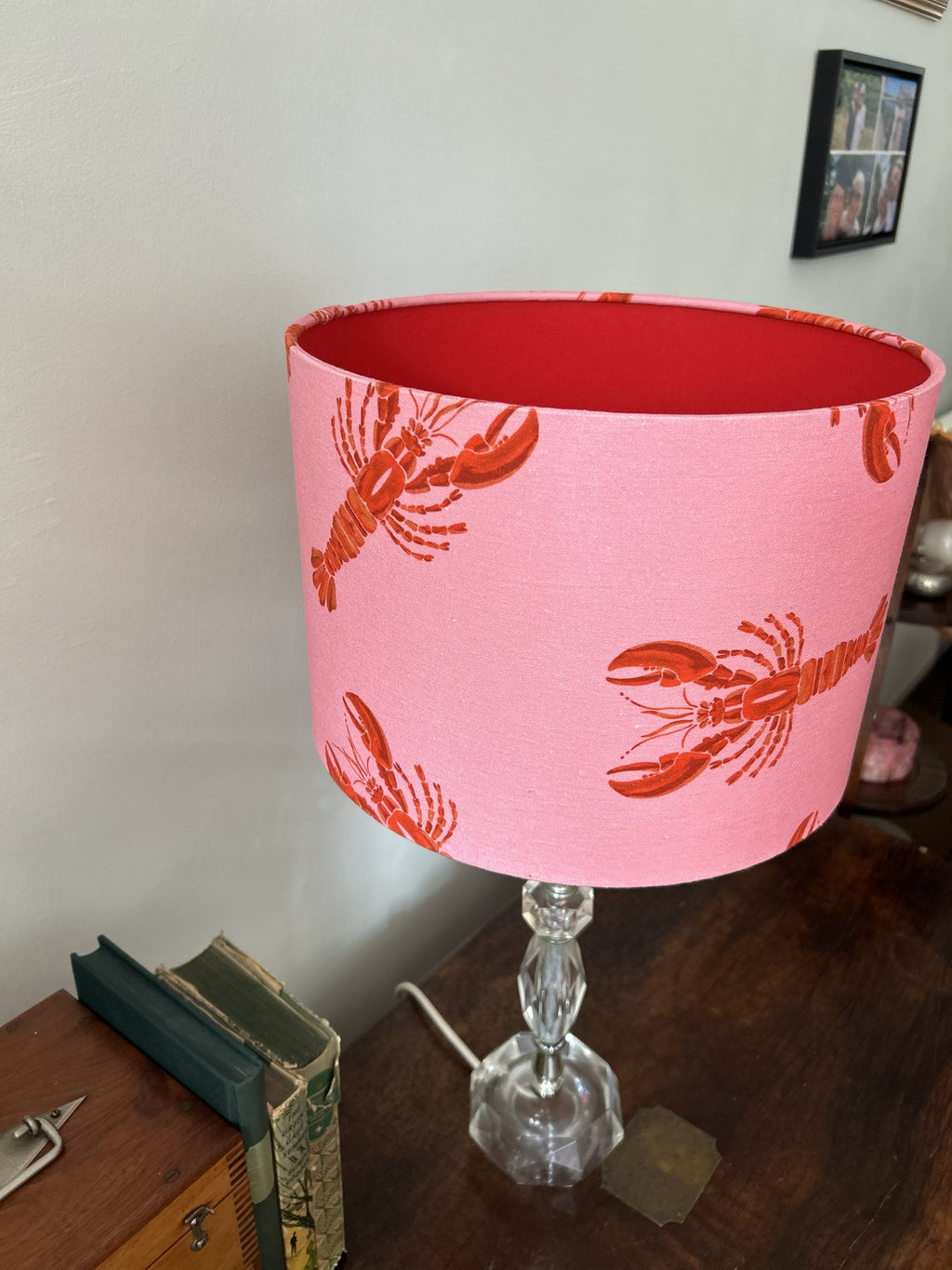 A pink fabric lampshade with a red lobster print, mounted on a clear glass table base and placed on a wooden table against a light wall.