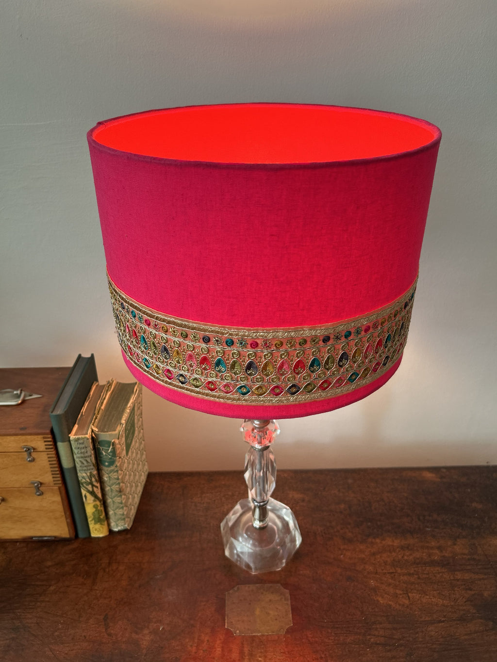 A vibrant magenta pink fabric lampshade with an ornate embroidered band featuring gold, red, and blue details, mounted on a clear faceted glass stem lamp, set on a wooden table.