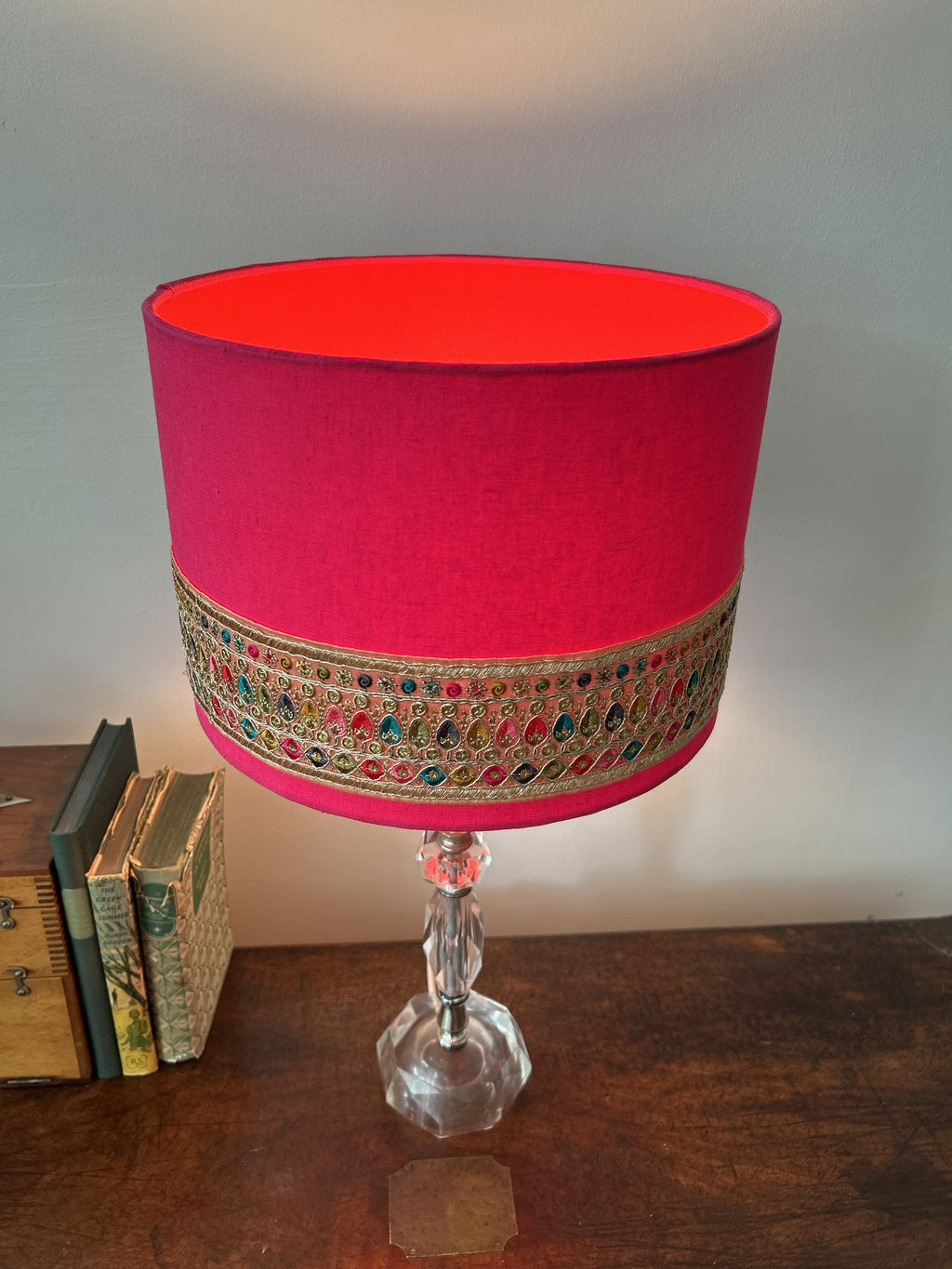 A vibrant magenta pink table lamp shade with an ornate embroidered band, mounted on a clear faceted glass pedestal base and set on a wooden table.