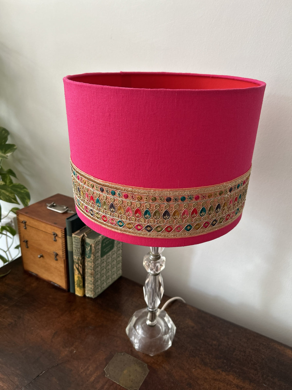 A bright pink fabric table lampshade with an ornate multicolor embroidered band around the middle, mounted on a clear glass pedestal base and shown on a wooden table.