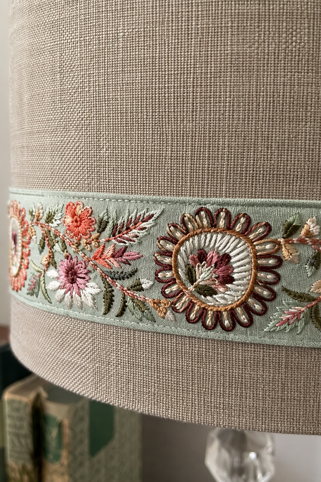 Close-up of a beige linen lampshade featuring a decorative embroidered band with floral and paisley motifs in muted green, pink, orange, and brown.