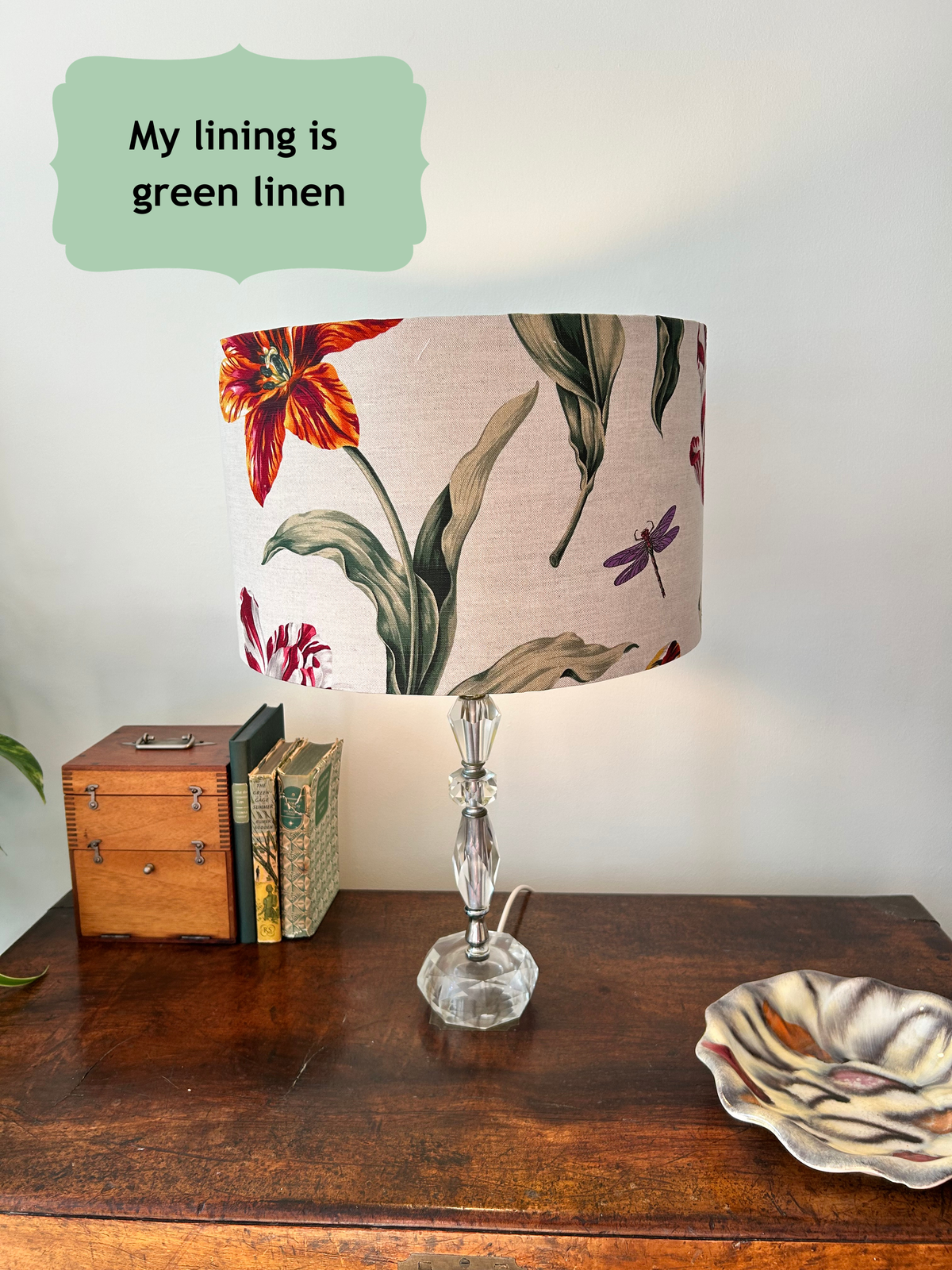 A table lamp with a large cylindrical drum shade featuring a botanical floral print in red, orange, green, and purple on a beige background, set on a dark wooden table next to vintage books and a ceramic dish. A light green text bubble in the top left reads 'My lining is green linen'.