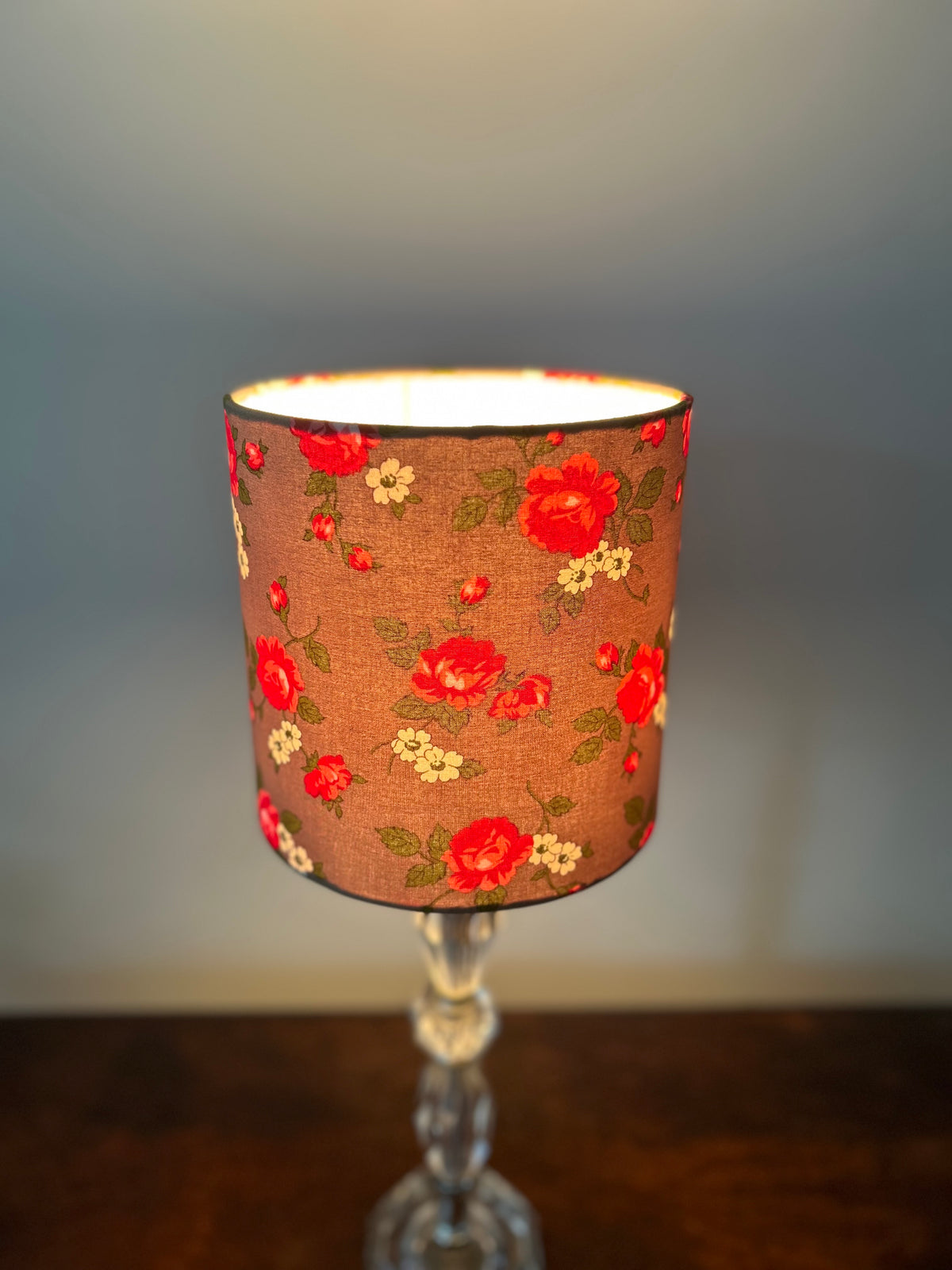 Lit Scattered Rosebud drum lampshade with red roses and small cream flowers on brown fabric, viewed slightly from above.