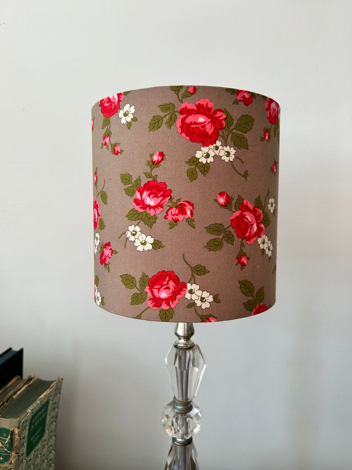 Unlit Scattered Rosebud drum lampshade with red roses, green leaves and cream flowers on taupe fabric, on a glass base.