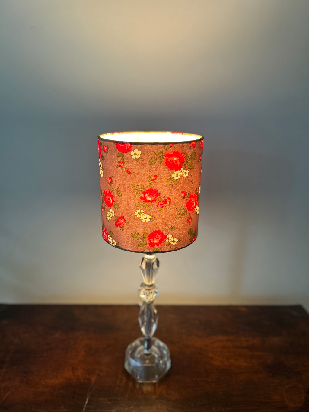 A table lamp with a cylindrical fabric shade featuring a red and yellow floral pattern on a warm brown background, mounted on a clear faceted glass stem and octagonal base, set on a dark wooden surface against a gray wall.