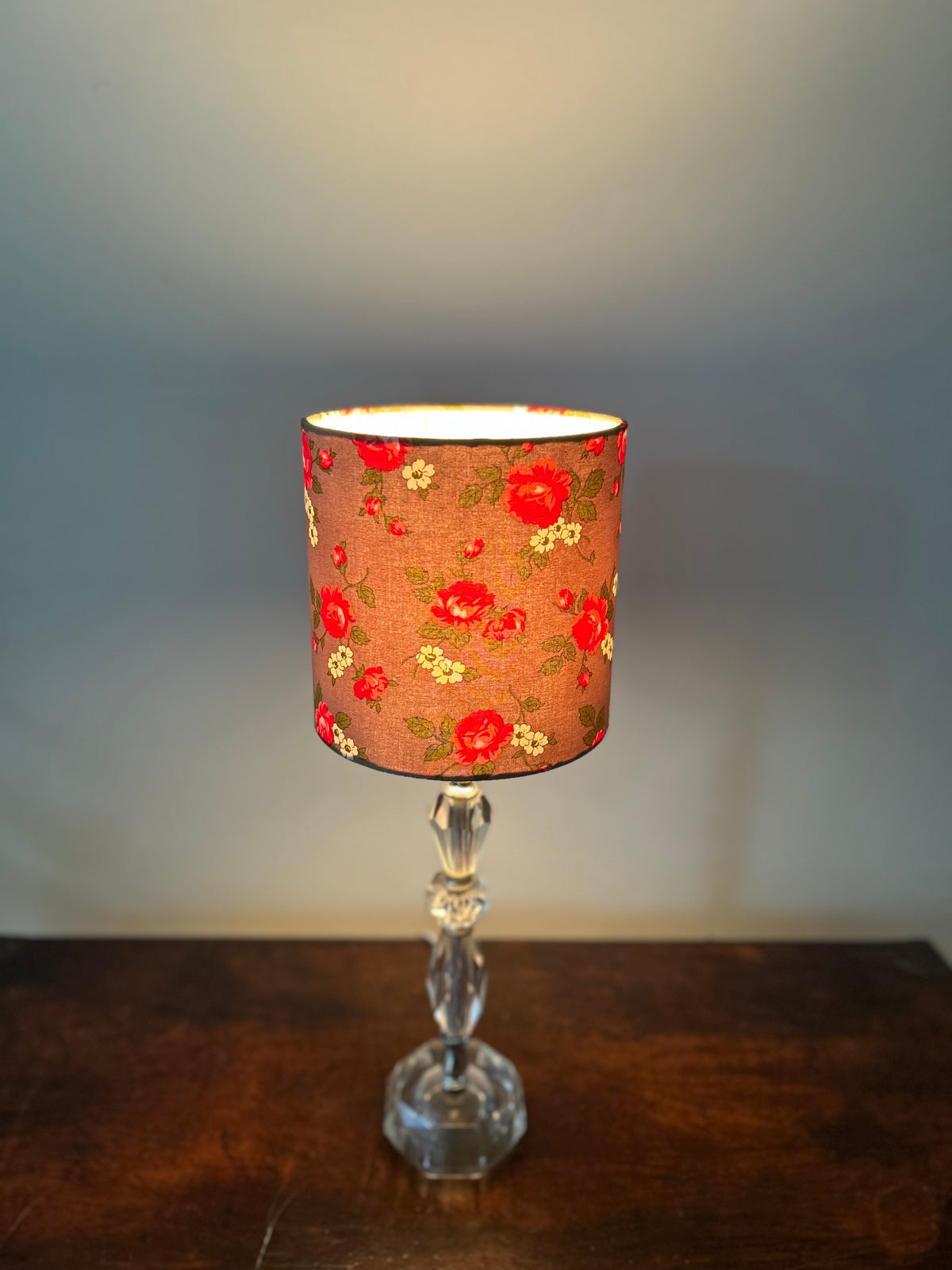 A table lamp with a cylindrical fabric shade featuring a red and yellow floral pattern on a warm brown background, mounted on a clear faceted glass stem and octagonal base, set on a dark wooden surface against a gray wall.