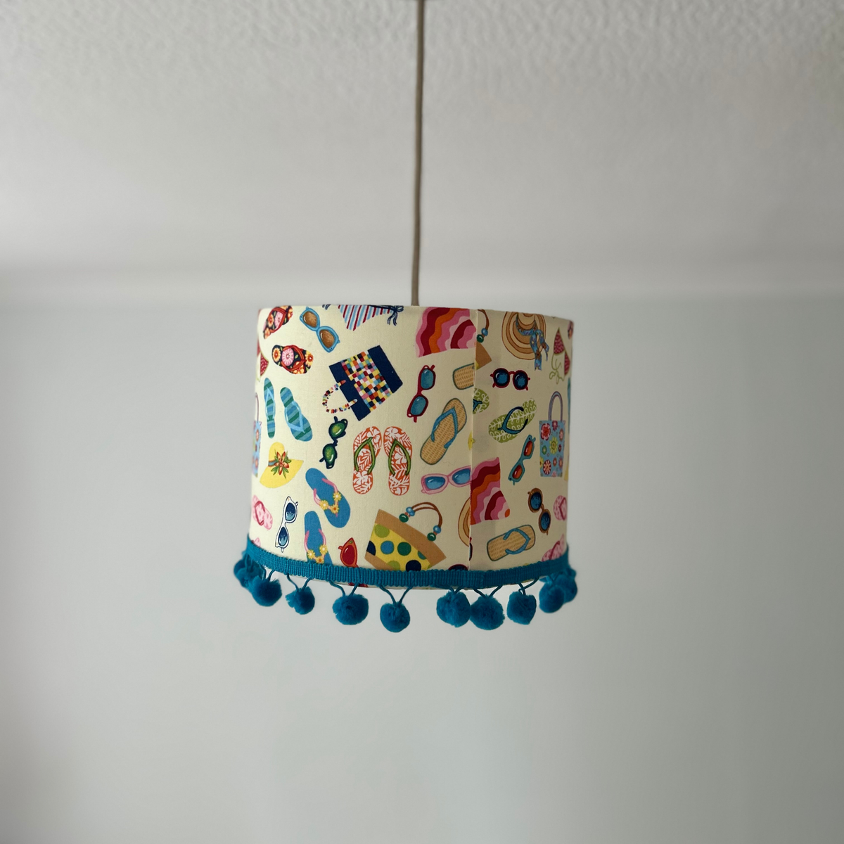 A cylindrical fabric lampshade with a colorful beach-themed print of flip-flops, umbrellas, and sunglasses, finished with a blue pom-pom trim at the bottom, hanging from a thin cord against a plain white wall.