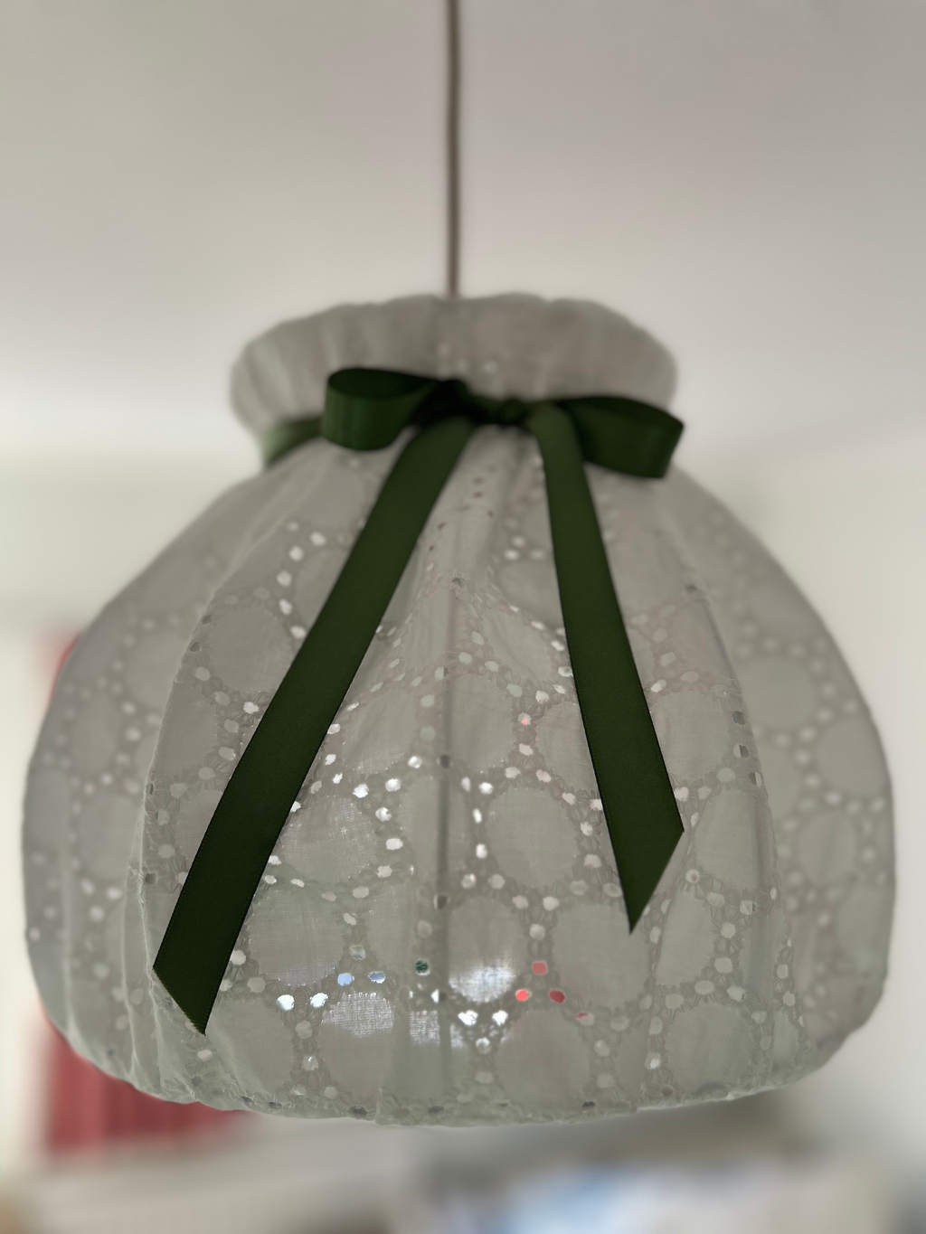 A white broiderie angliase pendant lampshade with a green ribbon bow, hanging against a neutral background.