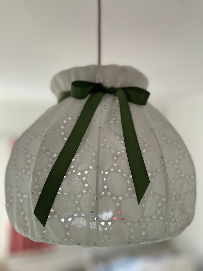A white broiderie angliase pendant lampshade with a green ribbon bow, hanging against a neutral background.