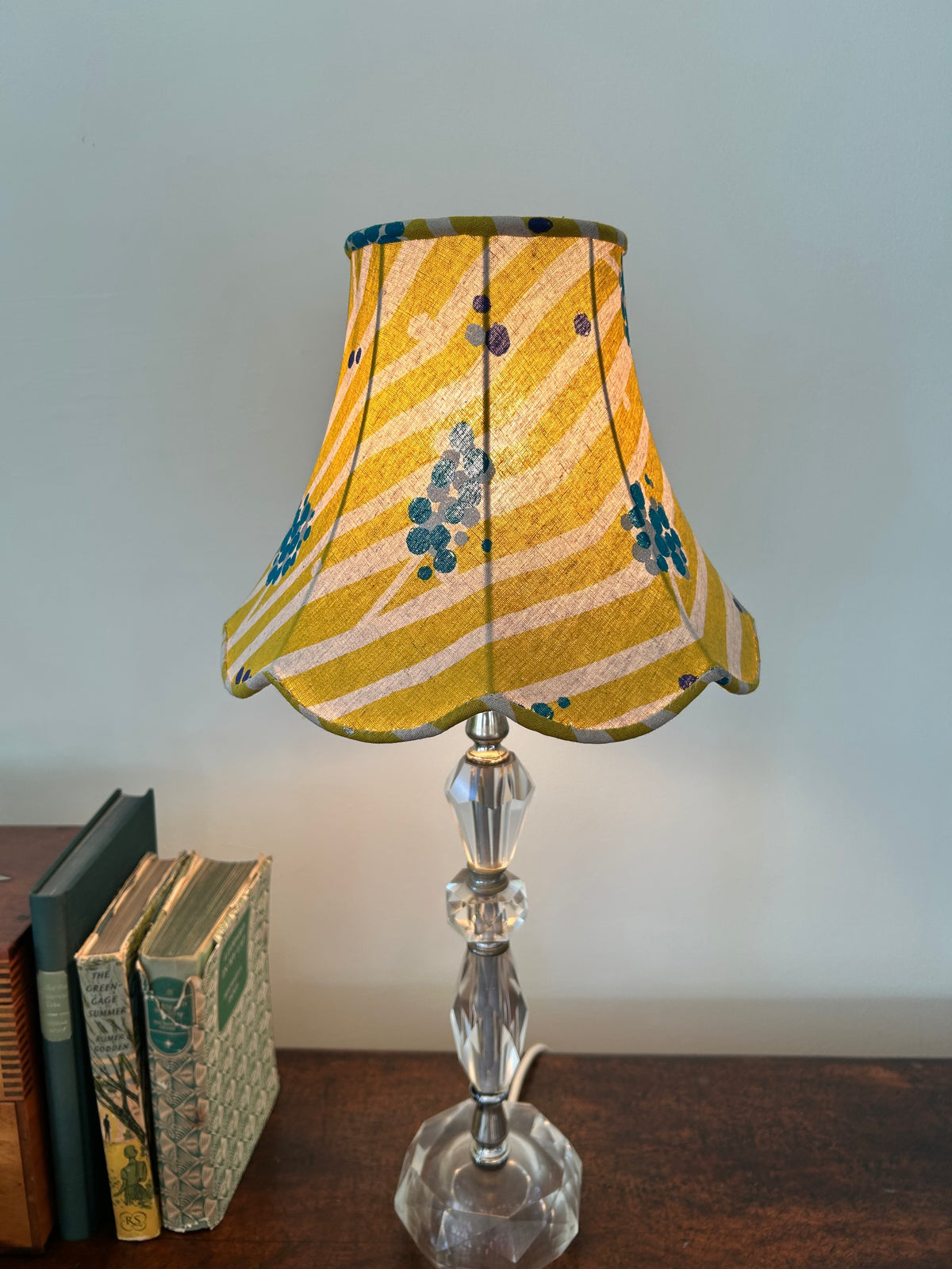 A table lamp with a lime and white striped fabric shade decorated with blue abstract floral motifs, mounted on a clear faceted glass stand, set on a wooden surface next to vintage books against a light gray wall.