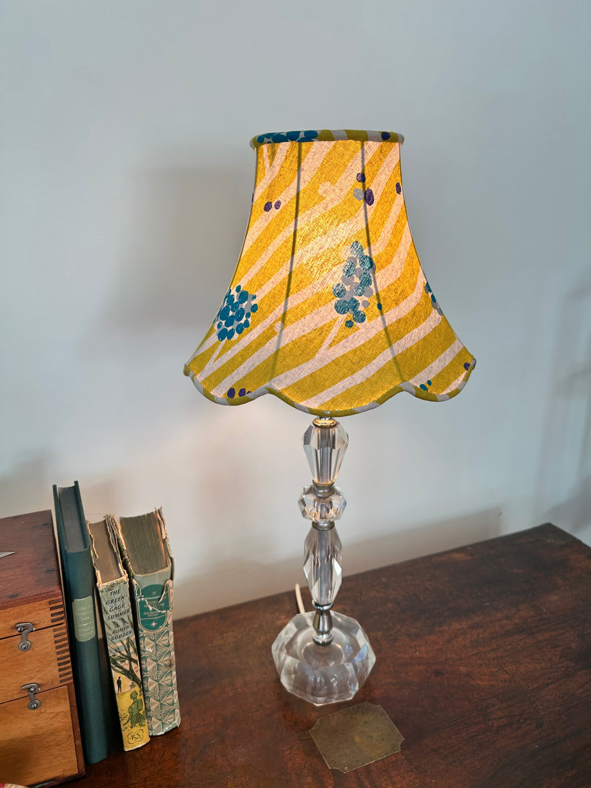 A vintage-style table lamp with a yellow and white striped fabric shade decorated with blue floral motifs, set on a dark wooden table next to old books against a plain wall.