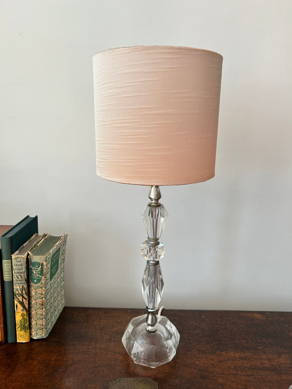 A table lamp with a tapered blush pink fabric shade and a clear faceted glass base, set on a dark wooden table next to a stack of vintage books against a plain light wall.
