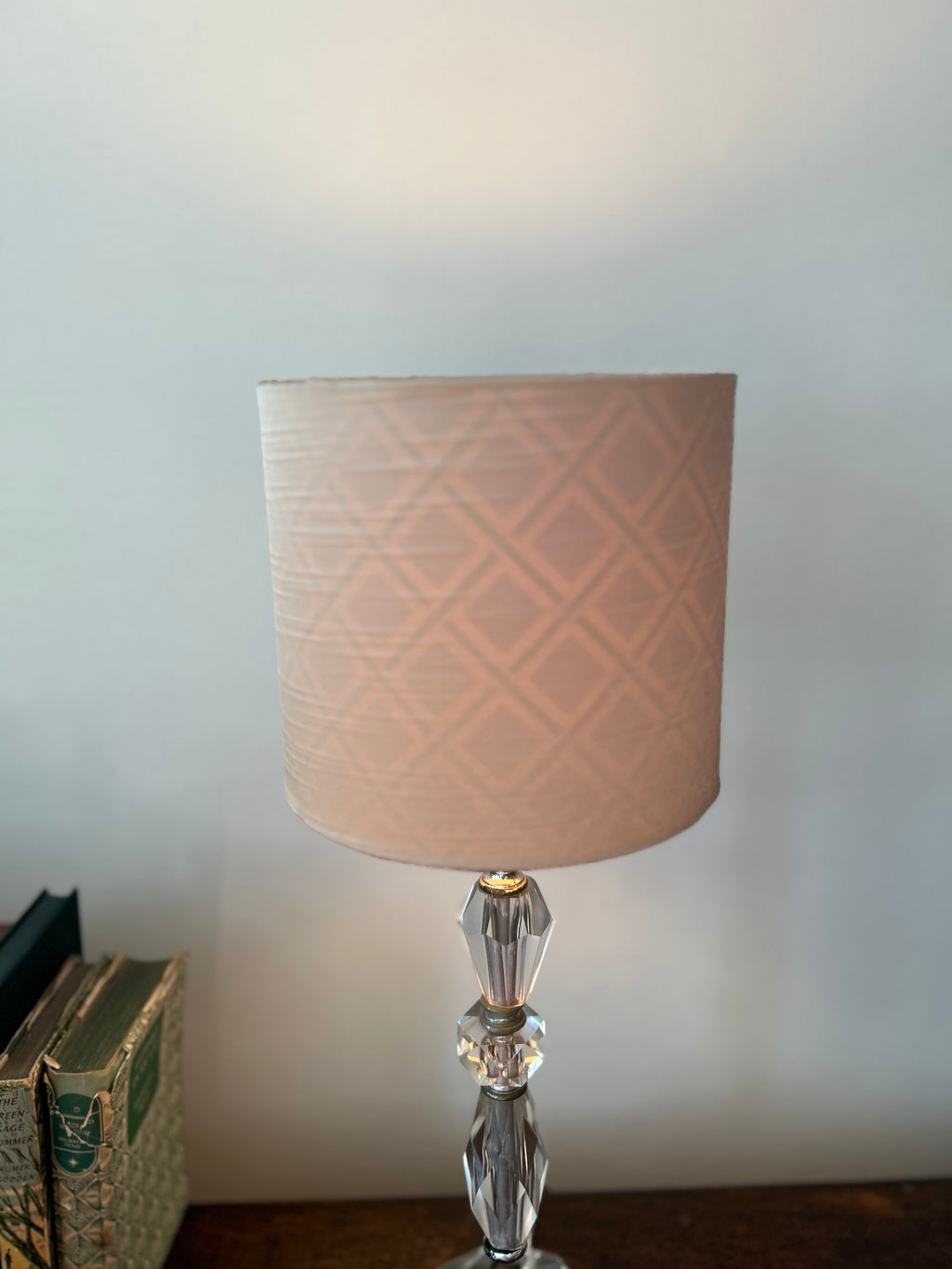 A table lamp with a light pink cylindrical shade featuring a subtle geometric diamond pattern shining through the blush satin, mounted on a faceted clear crystal base, set against a plain light gray wall.