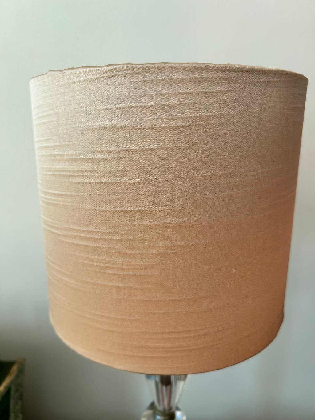 A beige fabric drum lampshade with horizontal pleats, shown on a table lamp with a clear glass base against a light gray wall.